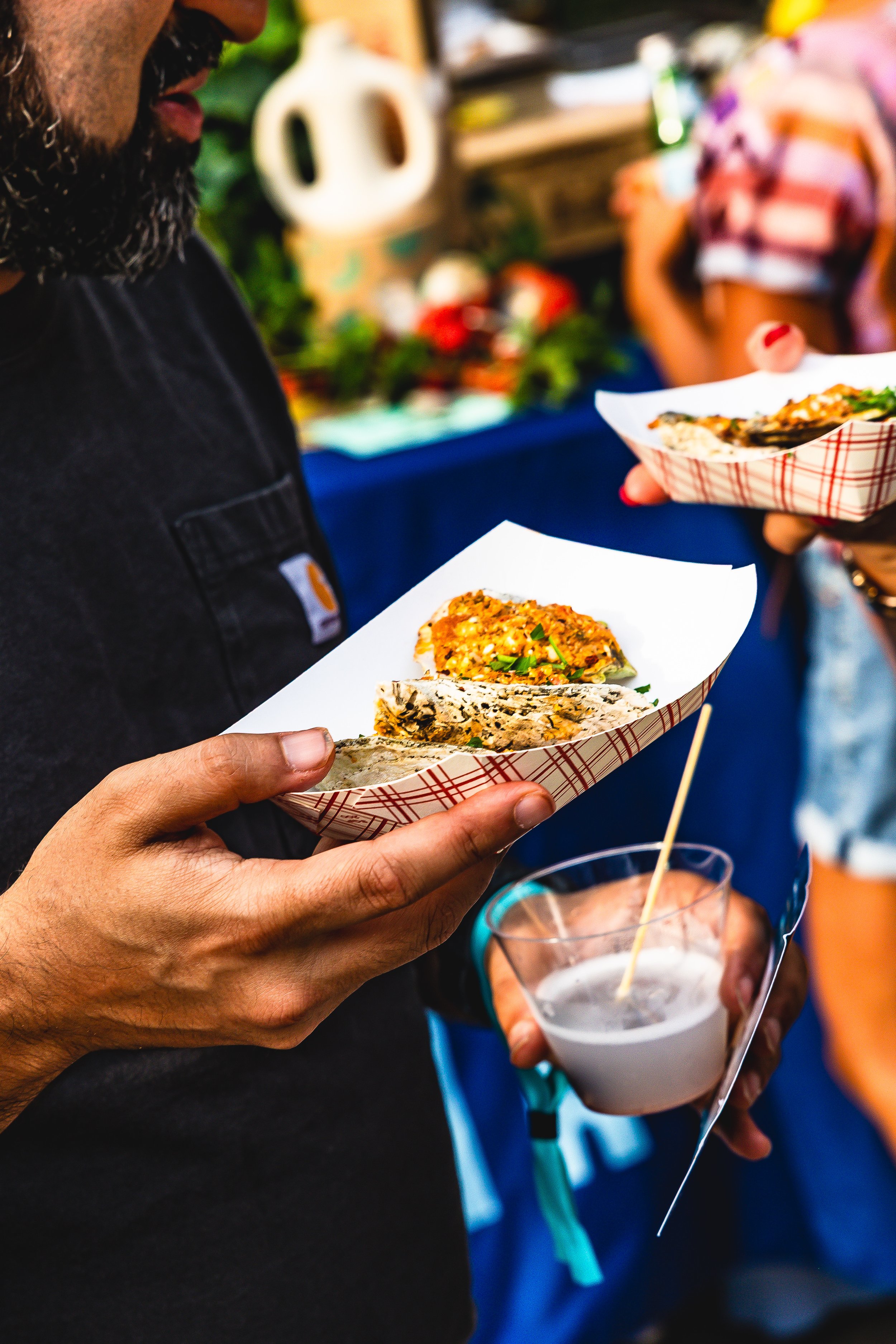 Person holding a paper tray with two tacos and a cup of drink at an outdoor food market.