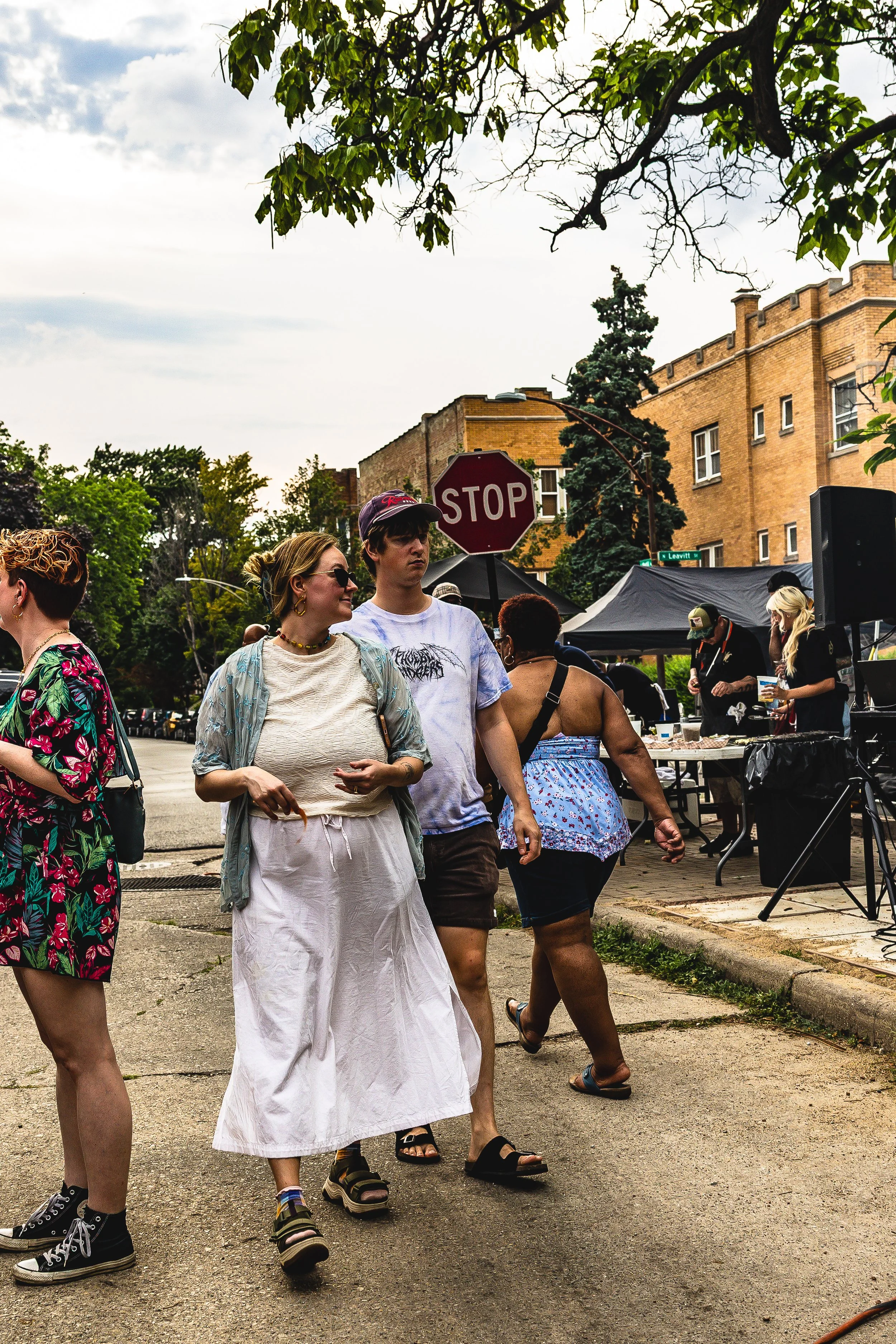 People walking on a sidewalk at an outdoor event with tents and a DJ, trees, a stop sign, and brick buildings in the background.