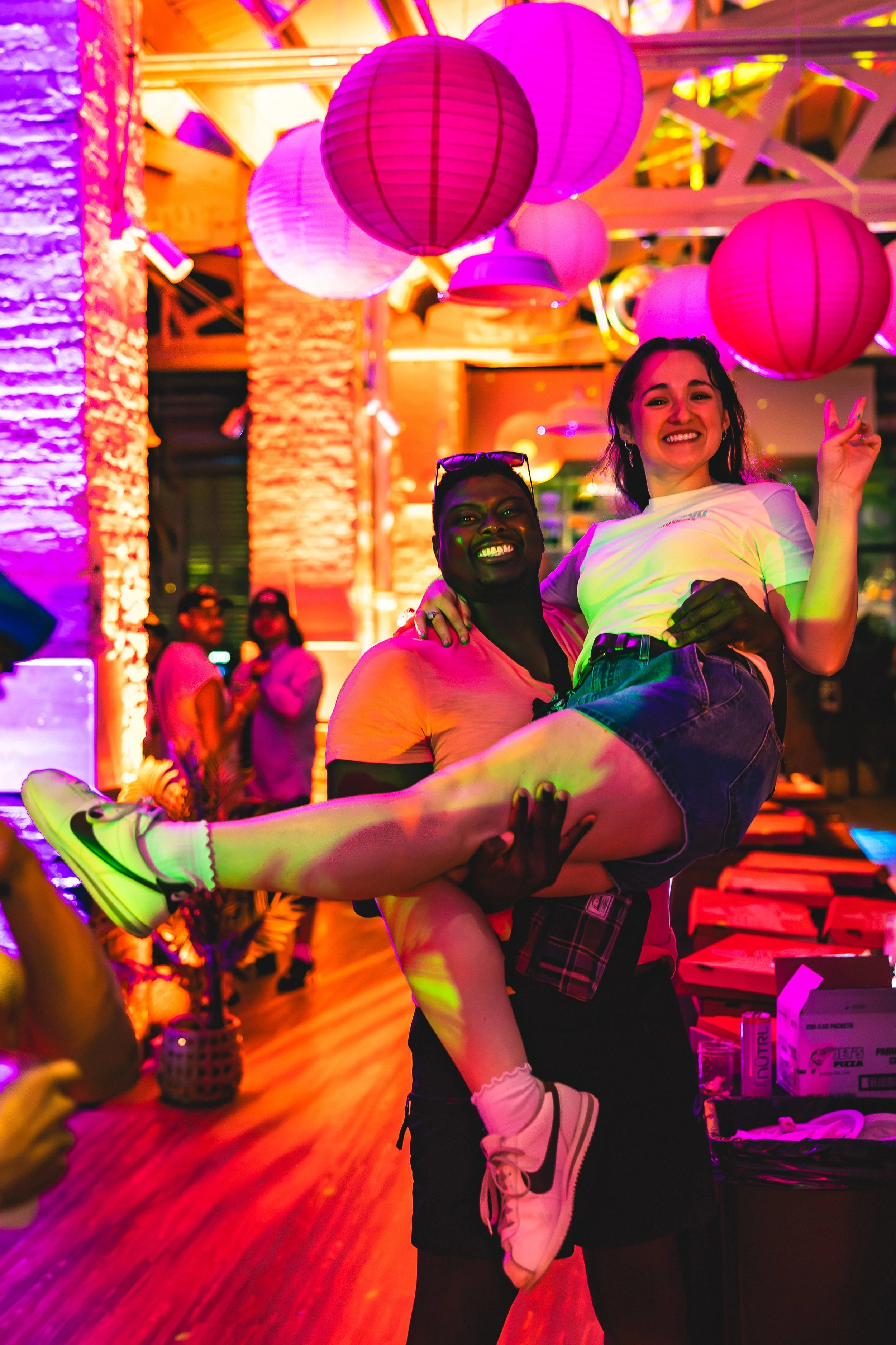 A person holding another person in a crowd at a party with colorful pink and purple lighting, lanterns, and decorations.