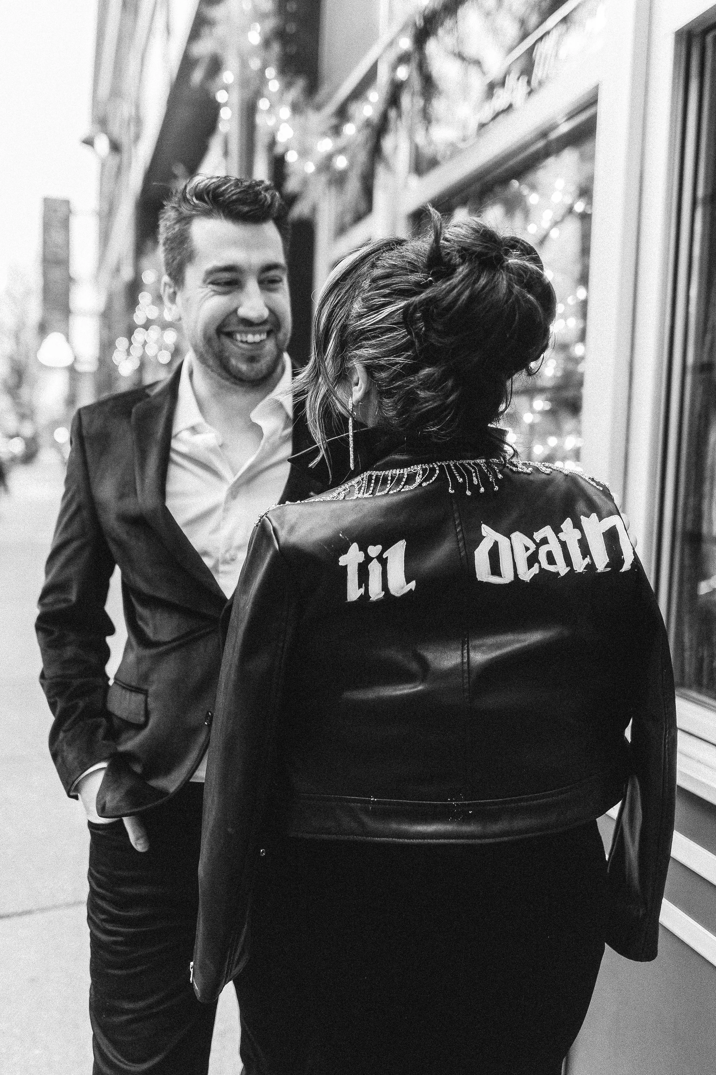 A man and a woman standing outside on a city street at night, smiling and talking to each other. The man is wearing a suit jacket and the woman is wearing a leather jacket with writing on the back. The background shows urban buildings and Christmas lights.