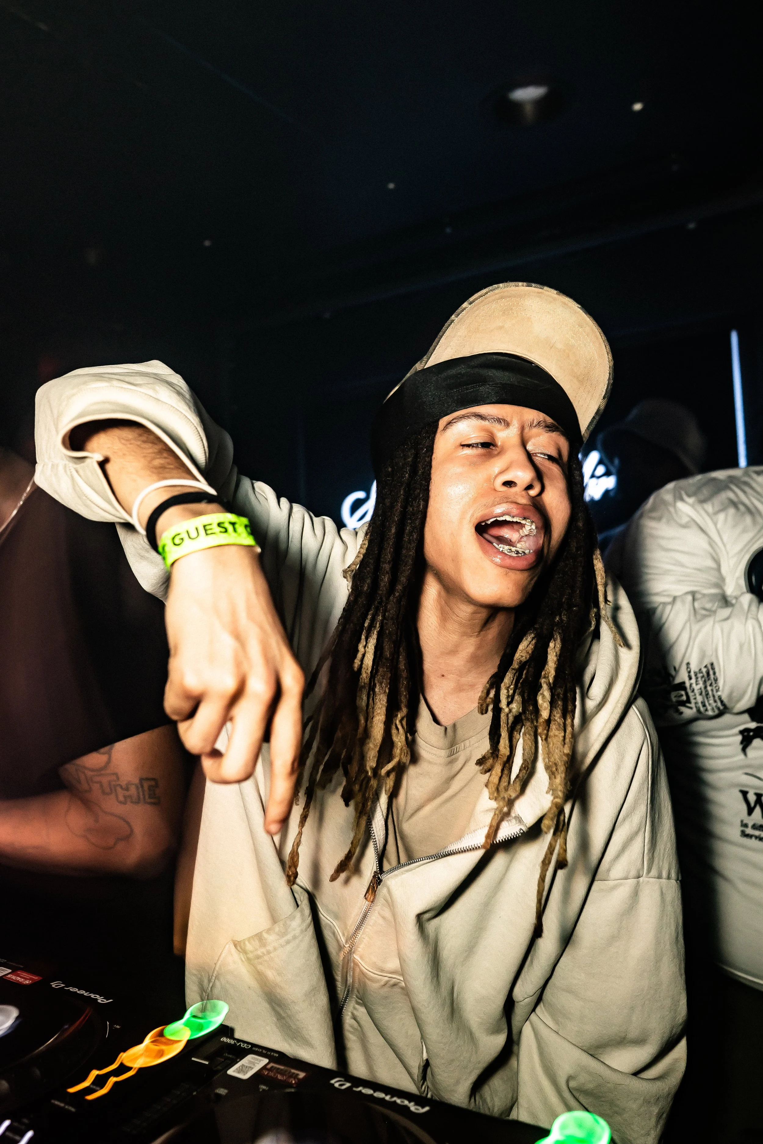 A DJ with dreadlocks wearing a baseball cap, headphones, and a zip-up hoodie, passionately mixing music at a nightclub with colorful lighting.