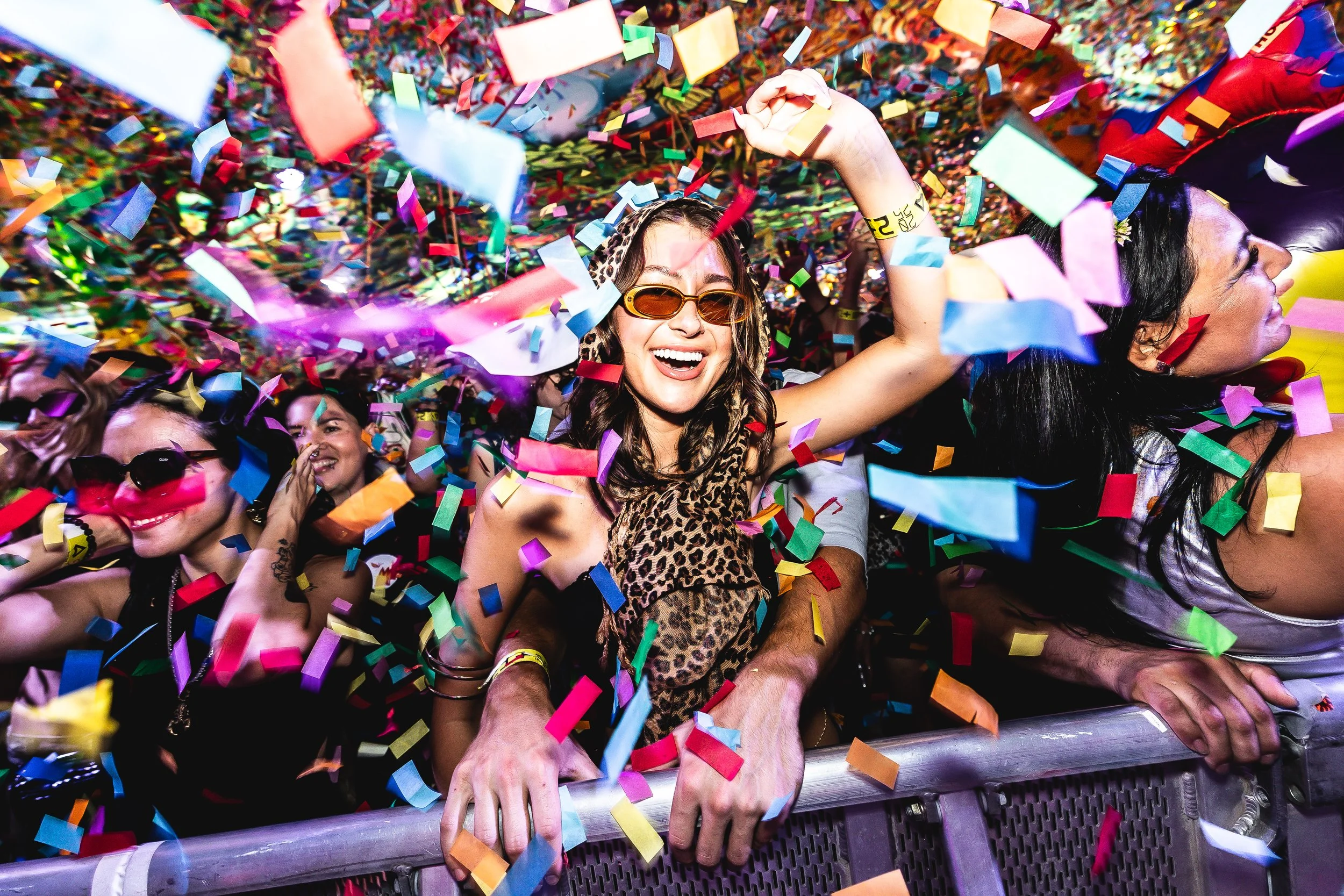 People celebrating at a party with colorful confetti falling around them, smiling and dancing near a barricade.
