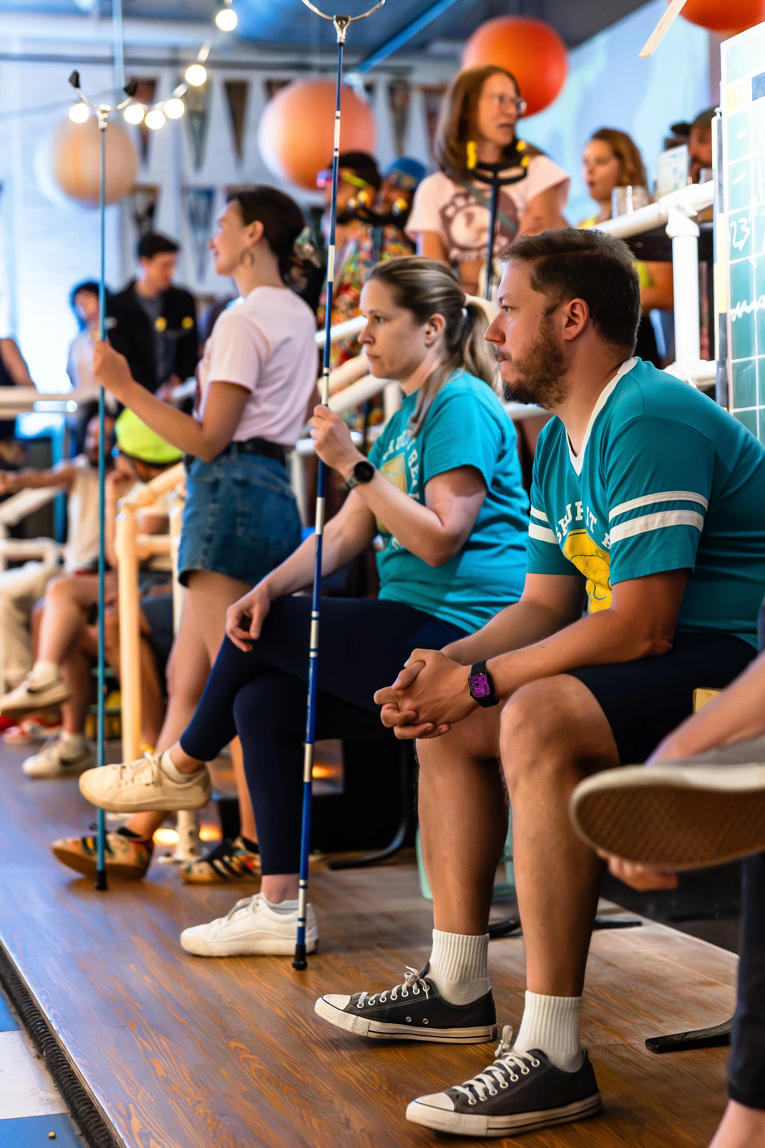 People sitting and standing on wooden bleachers at an outdoor event, some holding sticks with paper lanterns attached, under a blue canopy with string lights, with a chalkboard in the background.