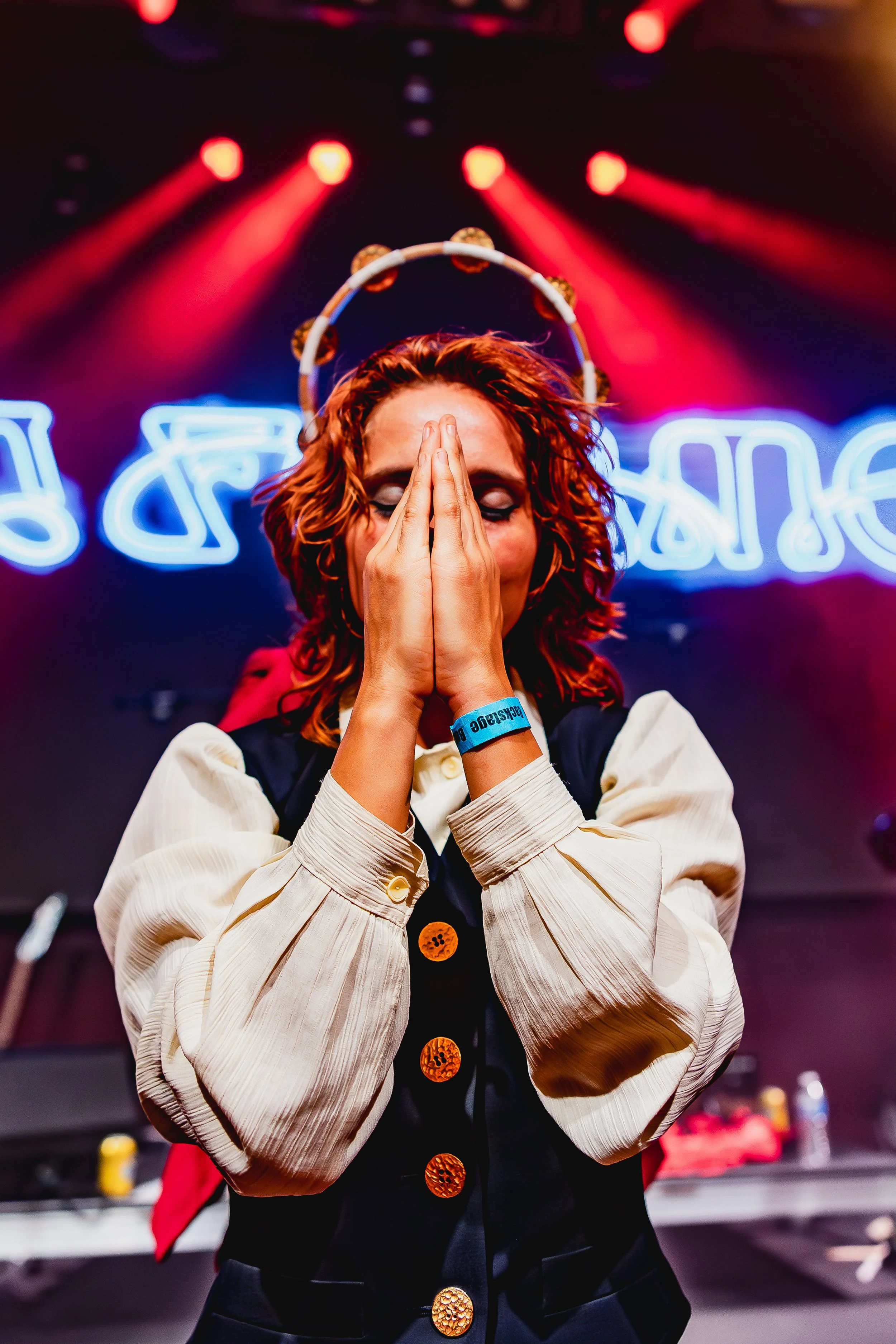 A woman with red curly hair and a headband, standing onstage with her hands pressed together in front of her face, in front of neon lights.