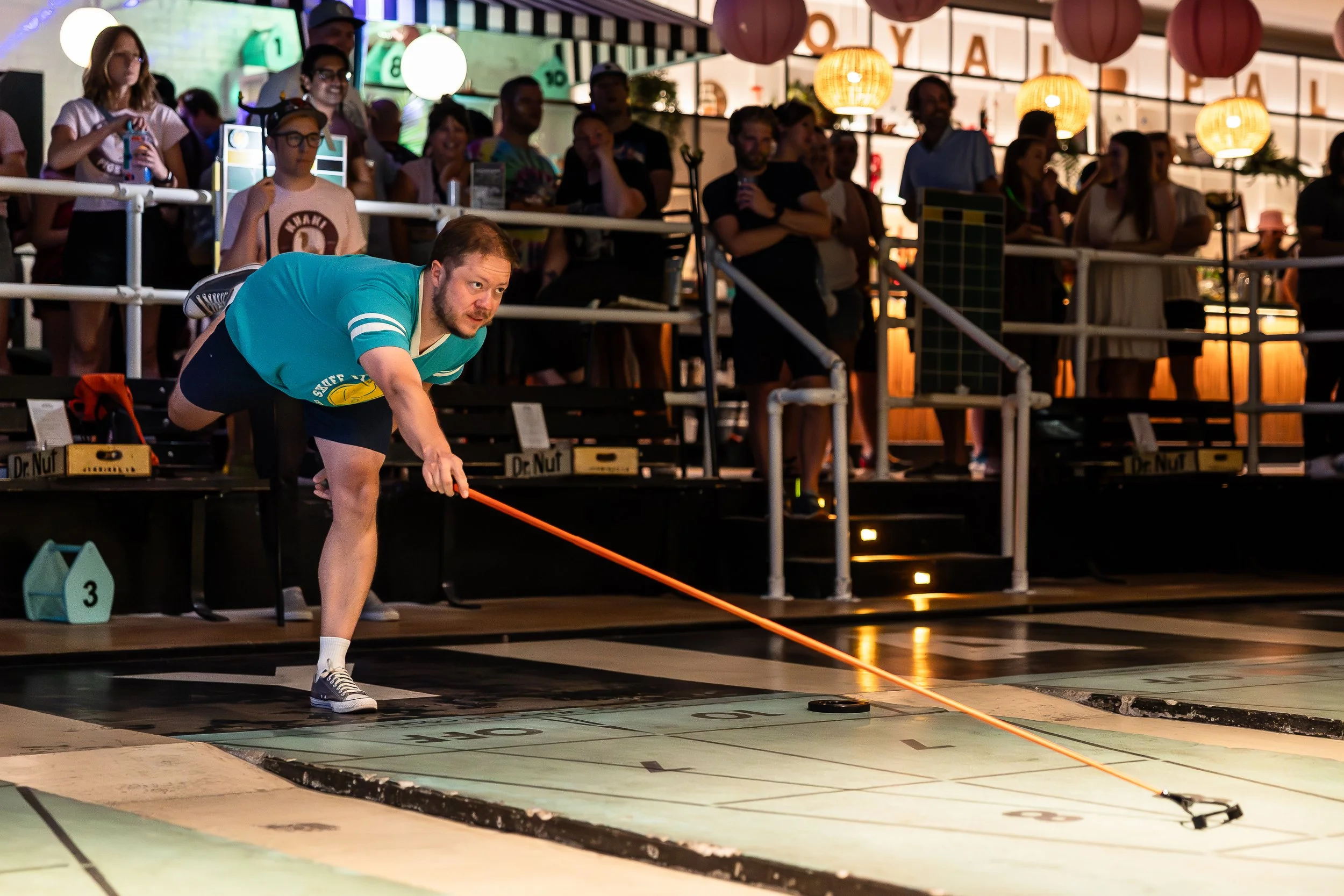 A man in a teal shirt and black shorts playing shuffleboard on a court with a crowd watching behind him at an indoor entertainment venue.