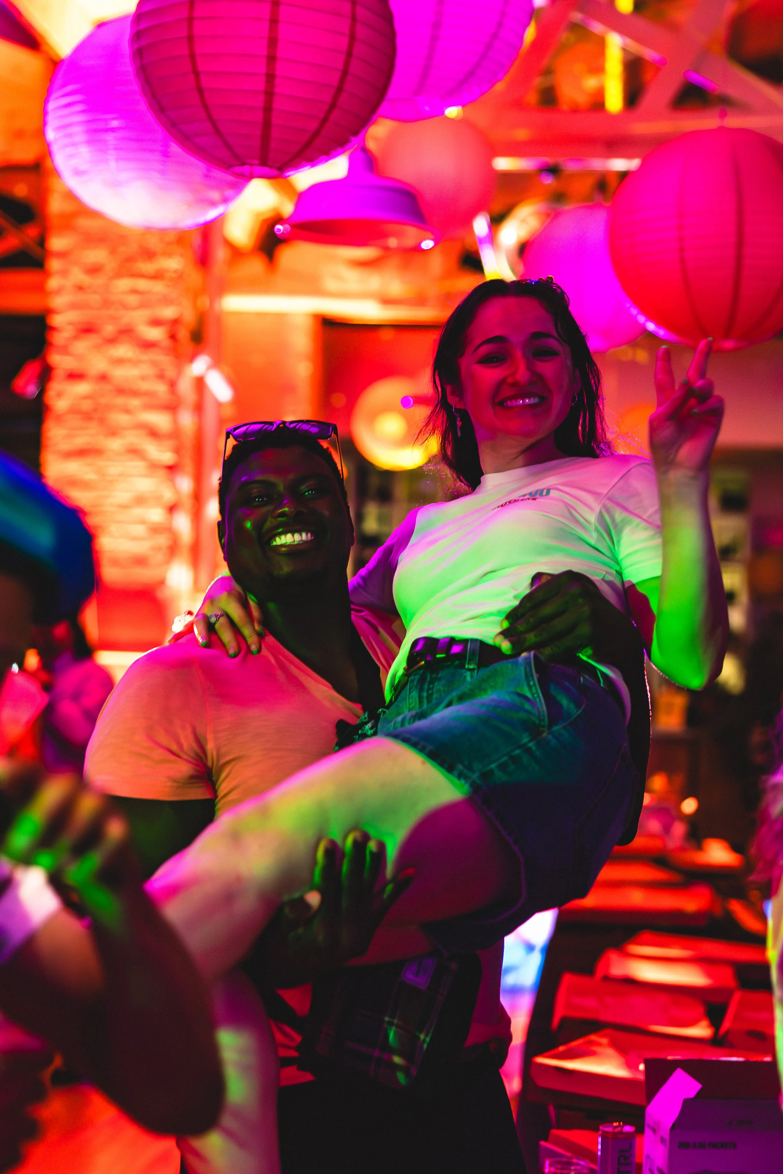 Two people in a colorful, lively setting with pink lanterns overhead, one person holding the other in a lift, both smiling.