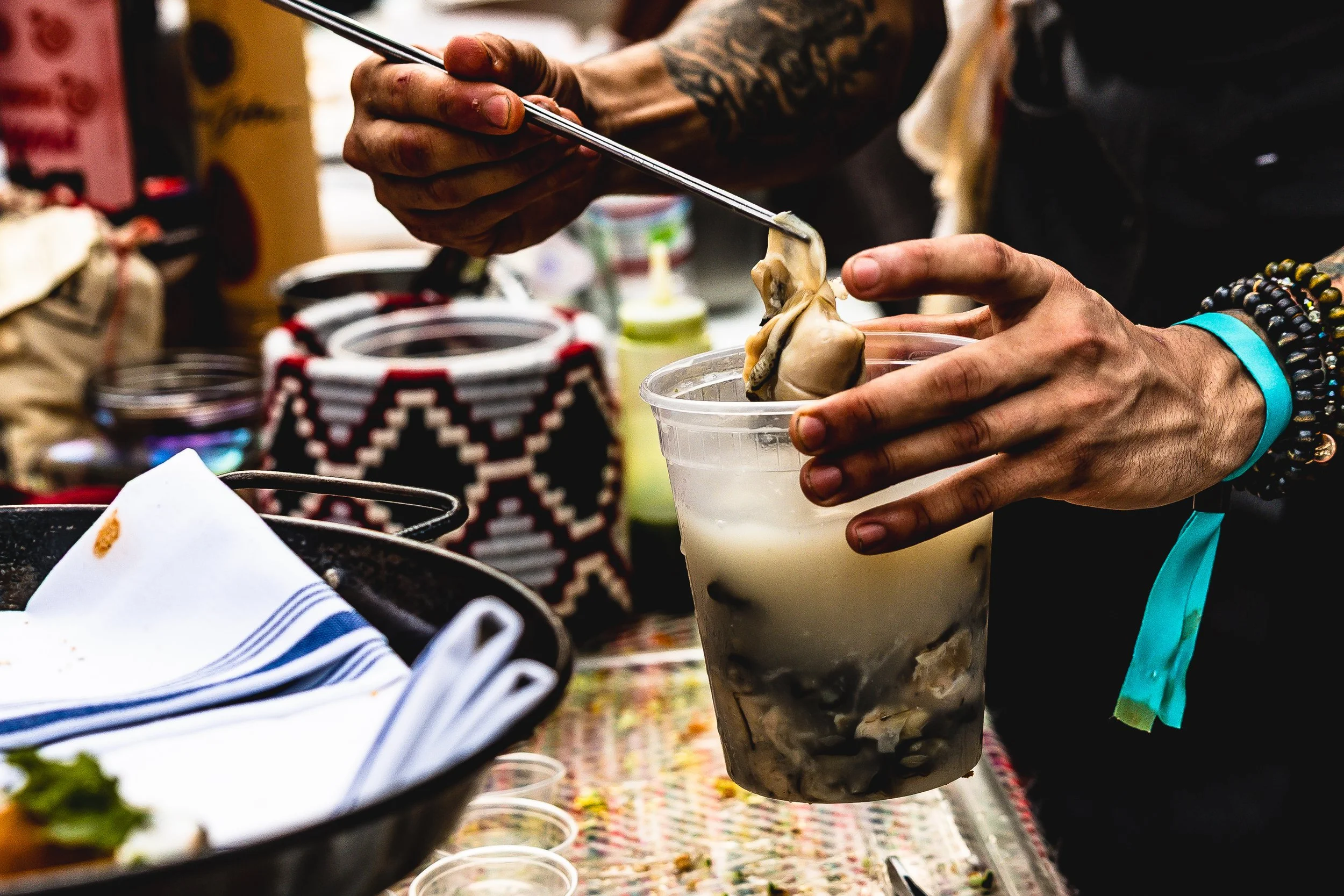 A person is inserting a plump oyster into a plastic cup of ice and liquid at a market stand. The person's hands have colorful bracelets, and the background includes various containers and items on a patterned cloth.
