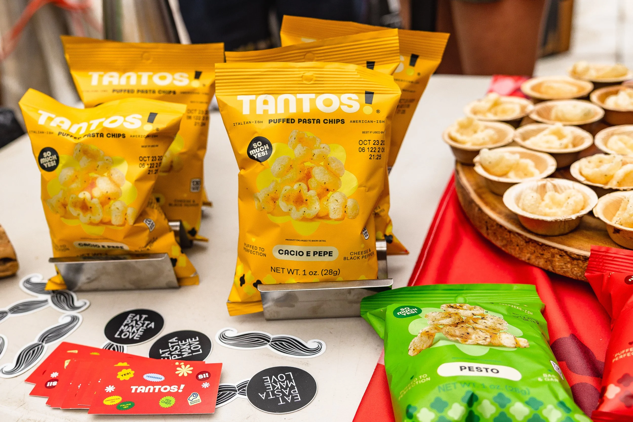 Bag of Tantos puffed pasta chips with cheese and black pepper flavor, lemon slices on the front, next to a green bag of Pesto flavored chips, and a tray of mini cups of a creamy dip or dessert on a red tablecloth.