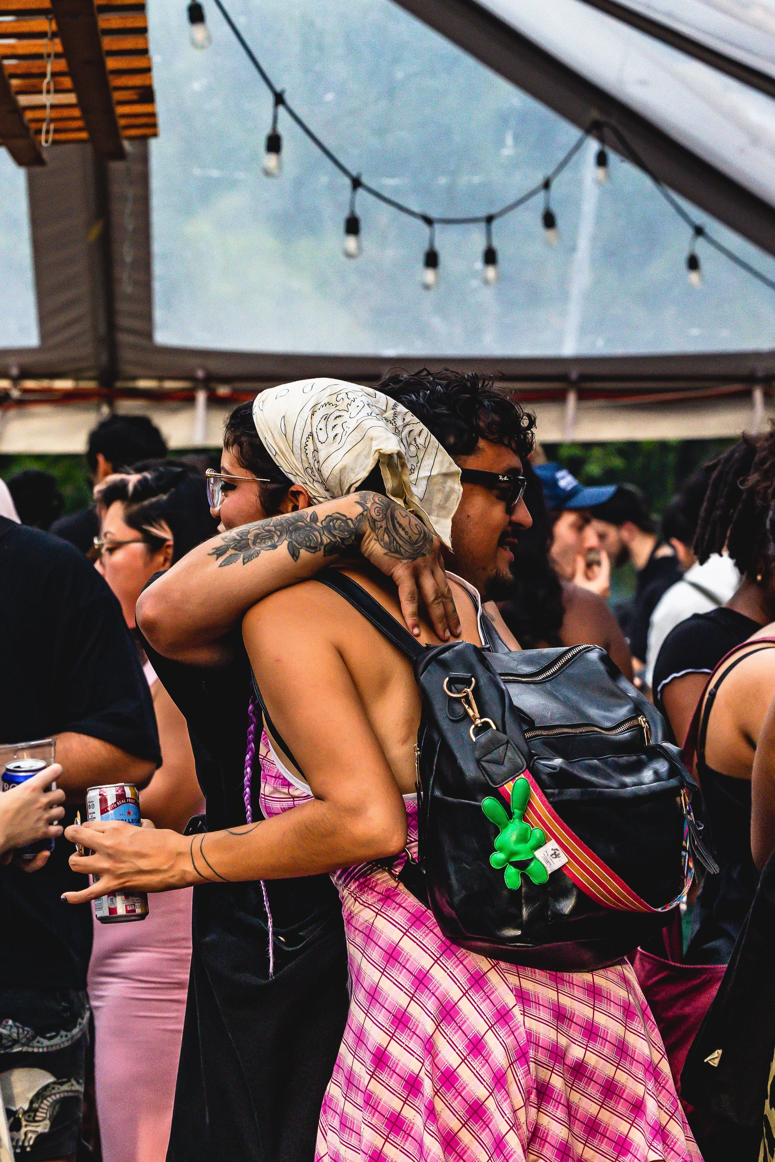 People at a crowded outdoor event, with one person hugging another, wearing sunglasses and a bandana, under a canopy with string lights.