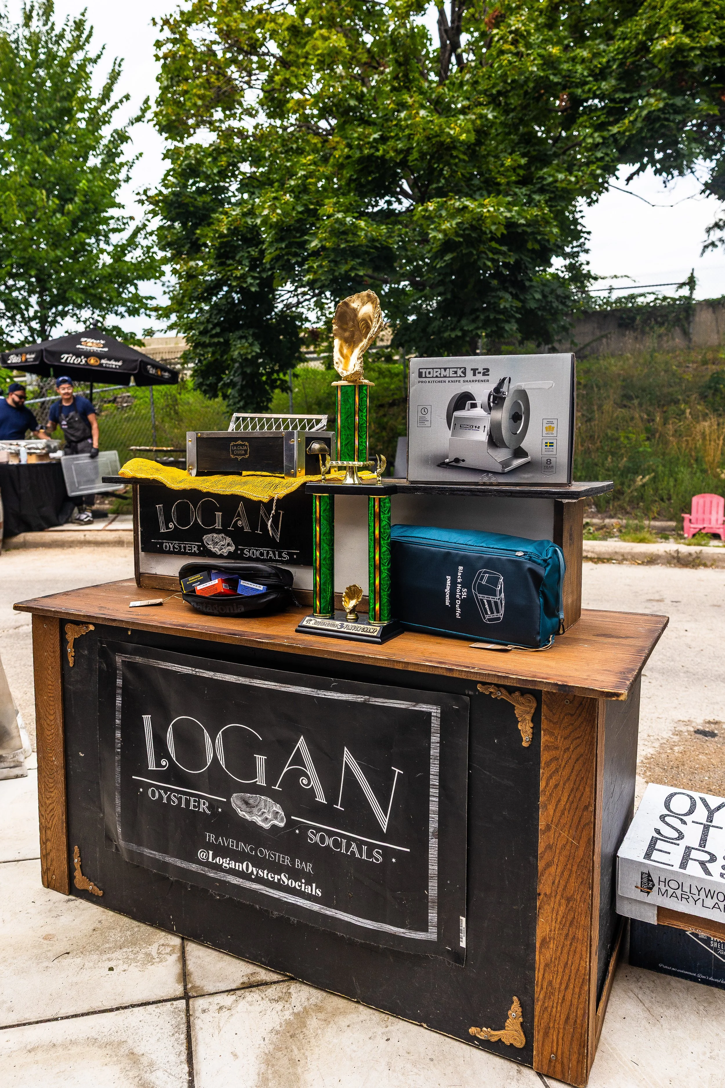Outdoor booth advertising Logan Oyster Socials with trophies, a knife sharpener, and various items on a wooden table, with trees and a fence in the background.