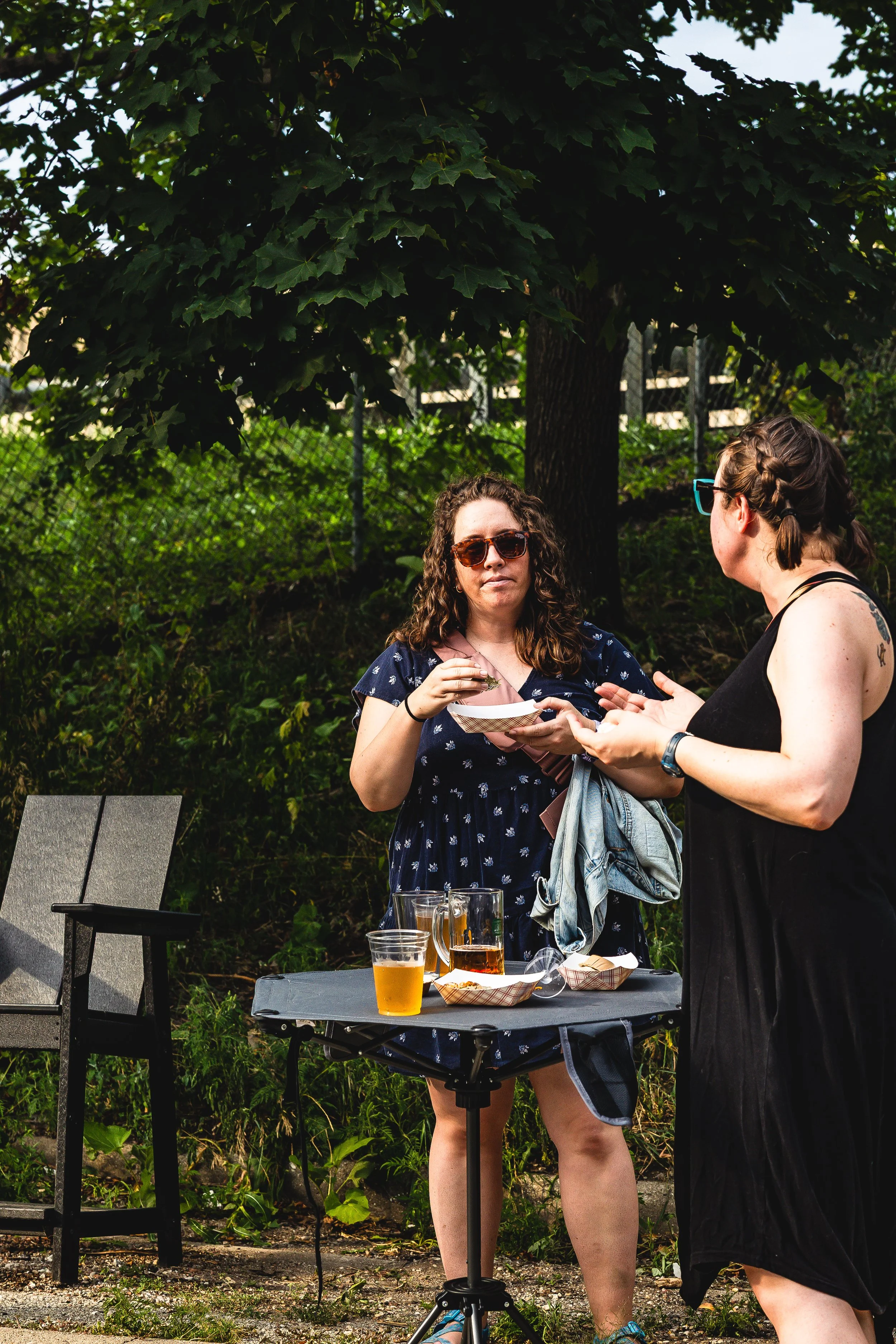 Two women are standing outdoors near a table with drinks, talking to each other. One woman has curly hair, wears sunglasses, and a navy dress. The other has braided hair, glasses, and a black dress. There are two empty chairs and a small table with f
