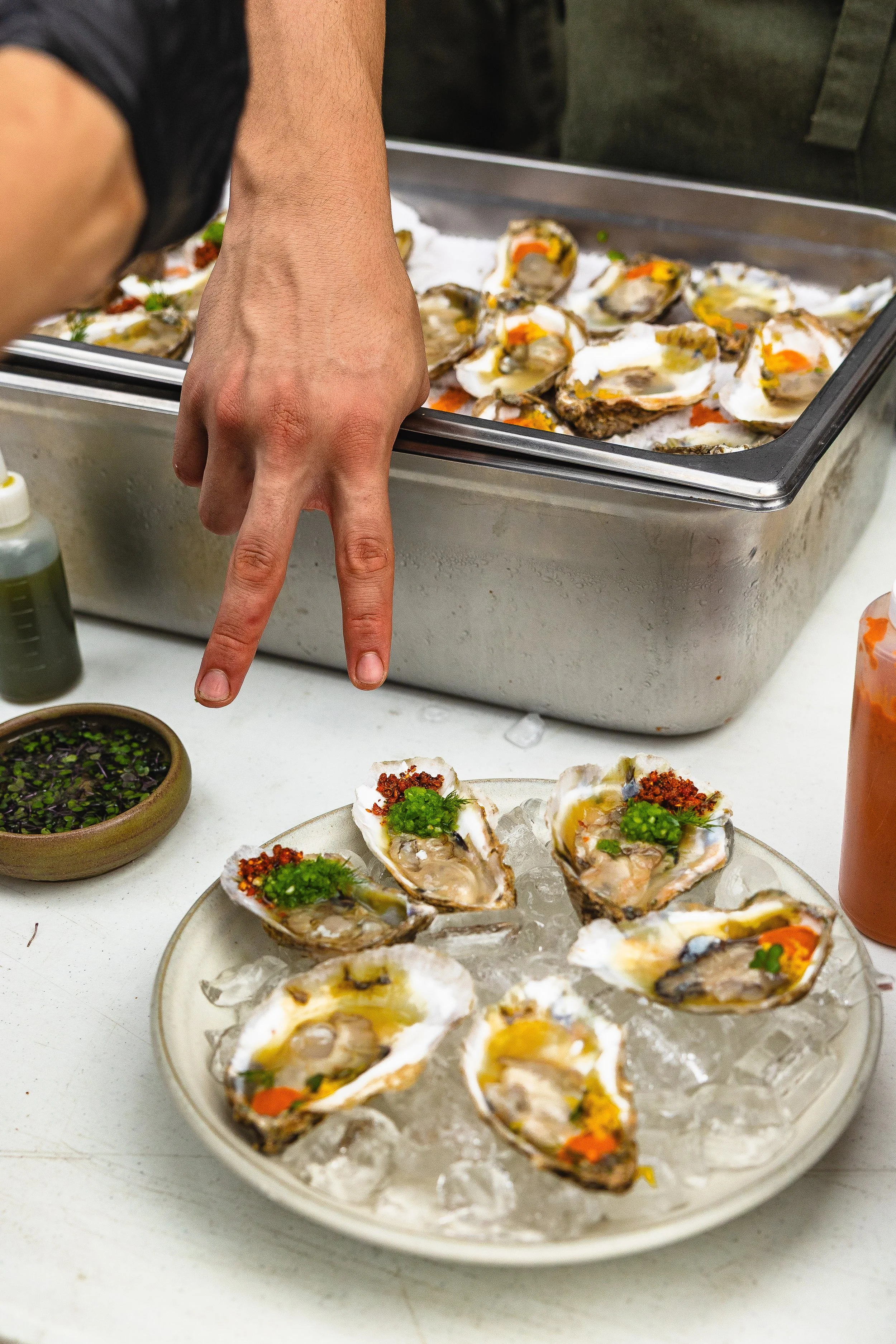 Close-up of a person preparing freshly shucked oysters on a bed of ice with hot sauce, green onions, and other toppings on a white plate, with a tray of oysters in the background.