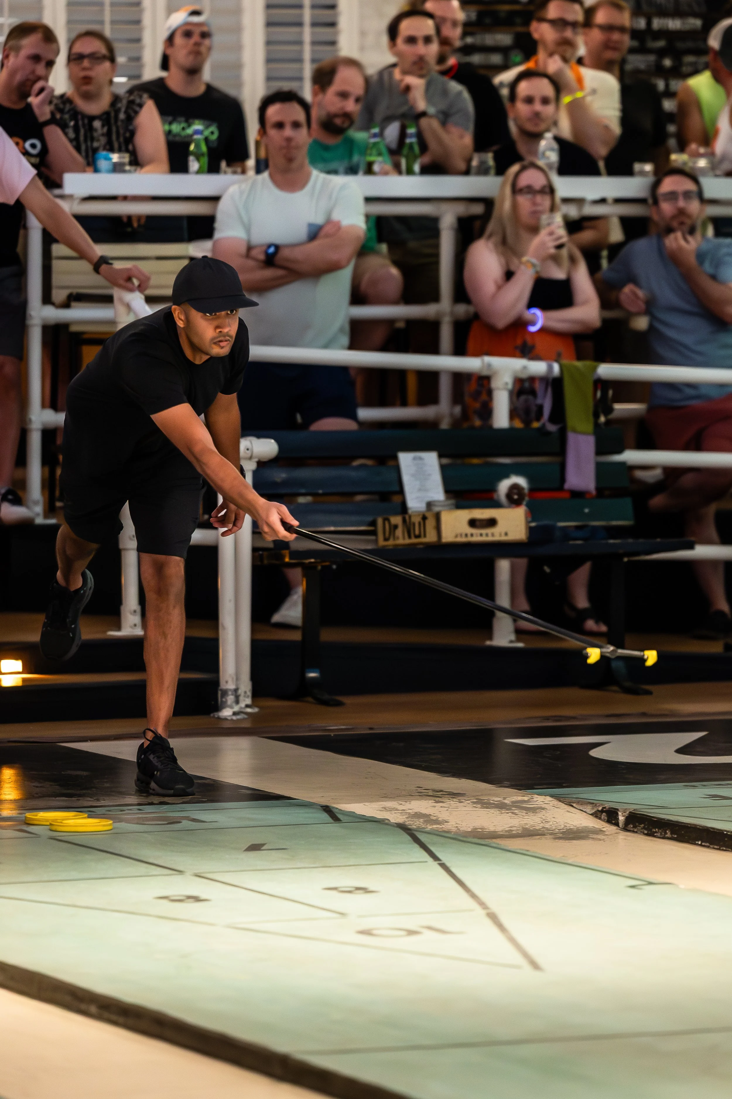 A man in a black shirt and shorts playing shuffleboard indoors while a group of spectators watches behind him.