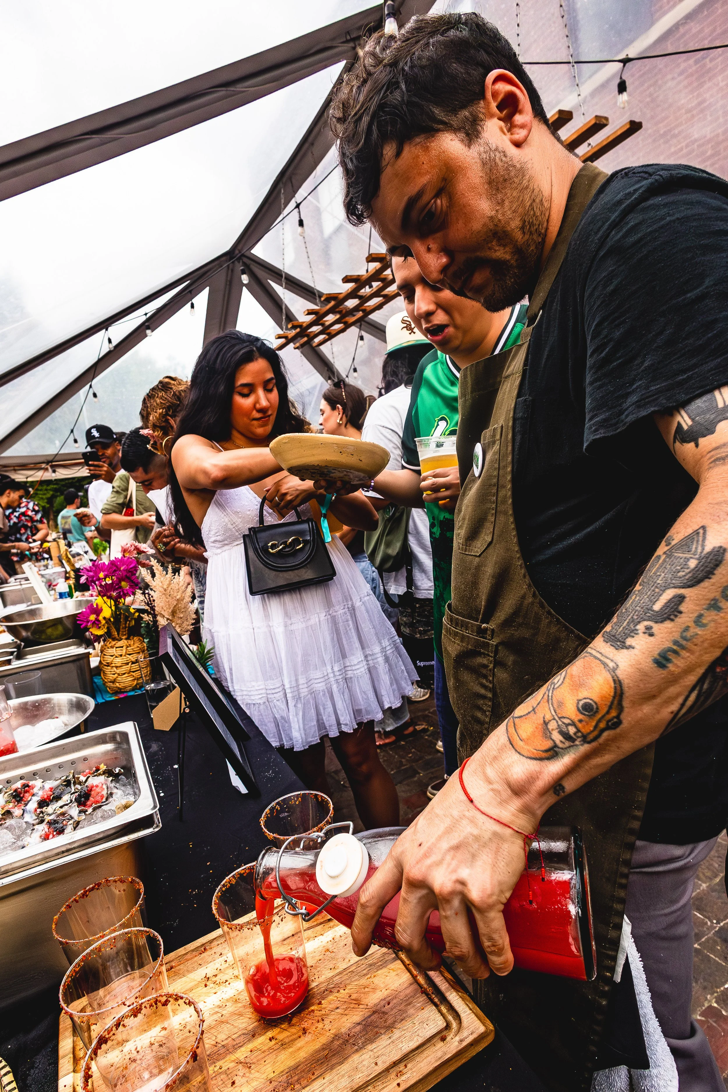 A group of people at a food event, with a man pouring red drink into glasses and others serving or selecting food from a table under a tent.