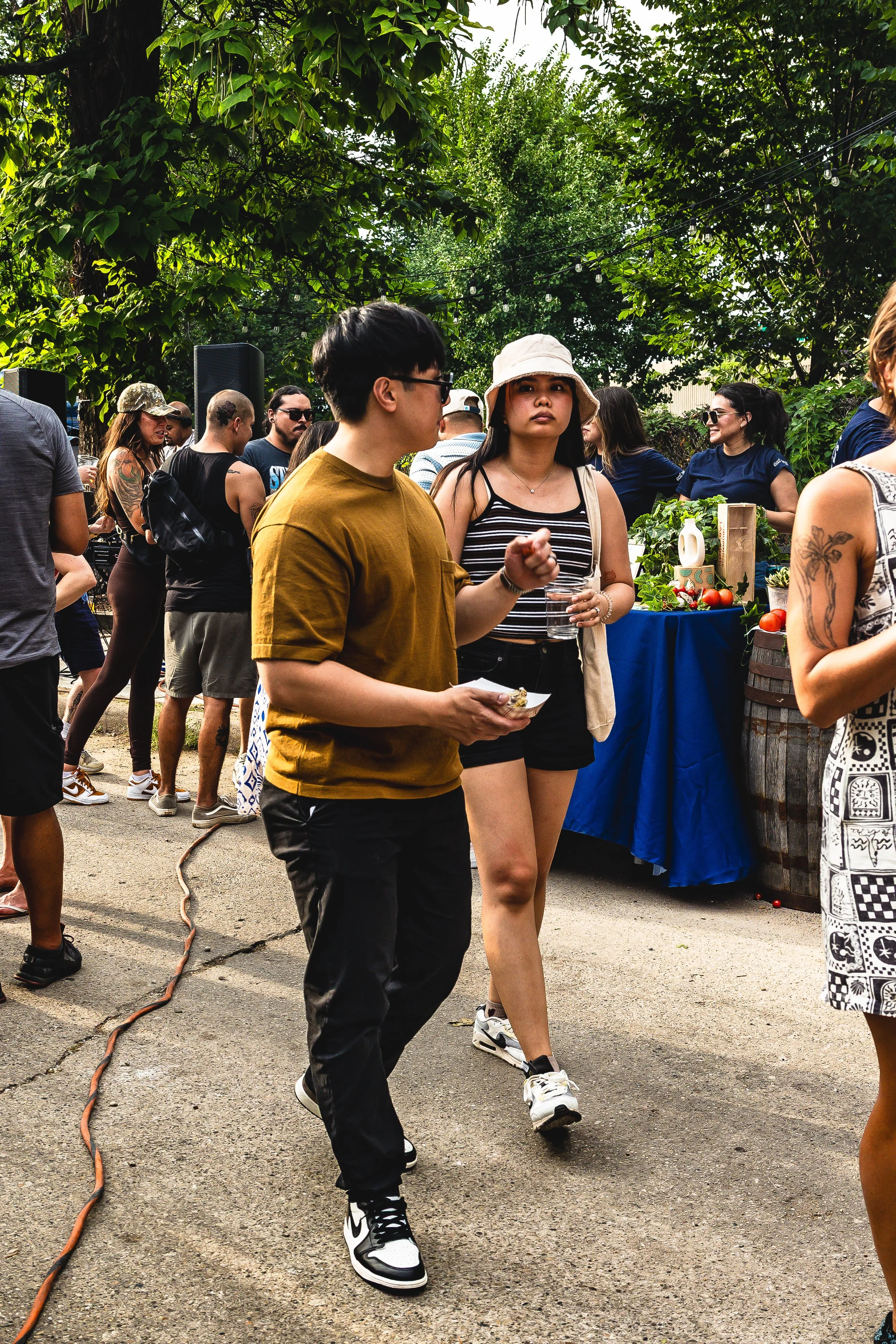 People attending an outdoor market or festival, with a woman in a striped top and bucket hat, and a man in a yellow shirt in the foreground, surrounded by greenery and vendor tables.