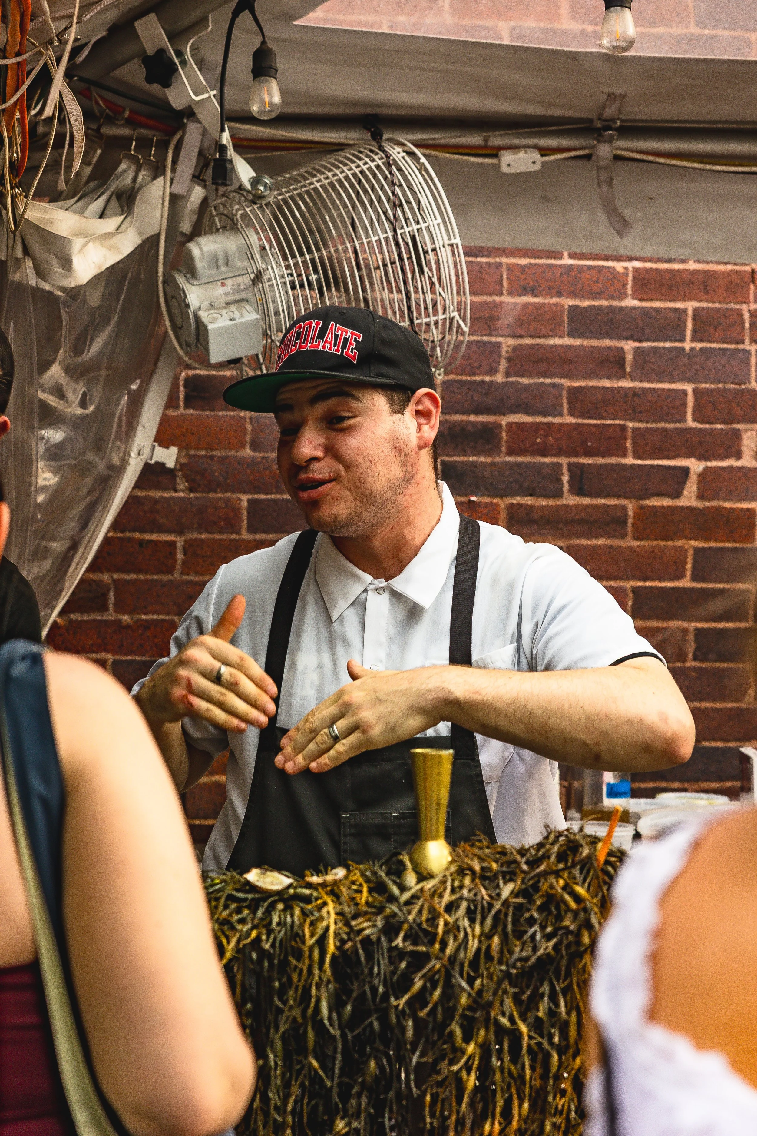 A man wearing a black baseball cap with 'CHOCOLATE' embroidered on it, a white shirt, and a black apron, standing behind a booth decorated with dried plants, speaking to customers against a brick wall background.