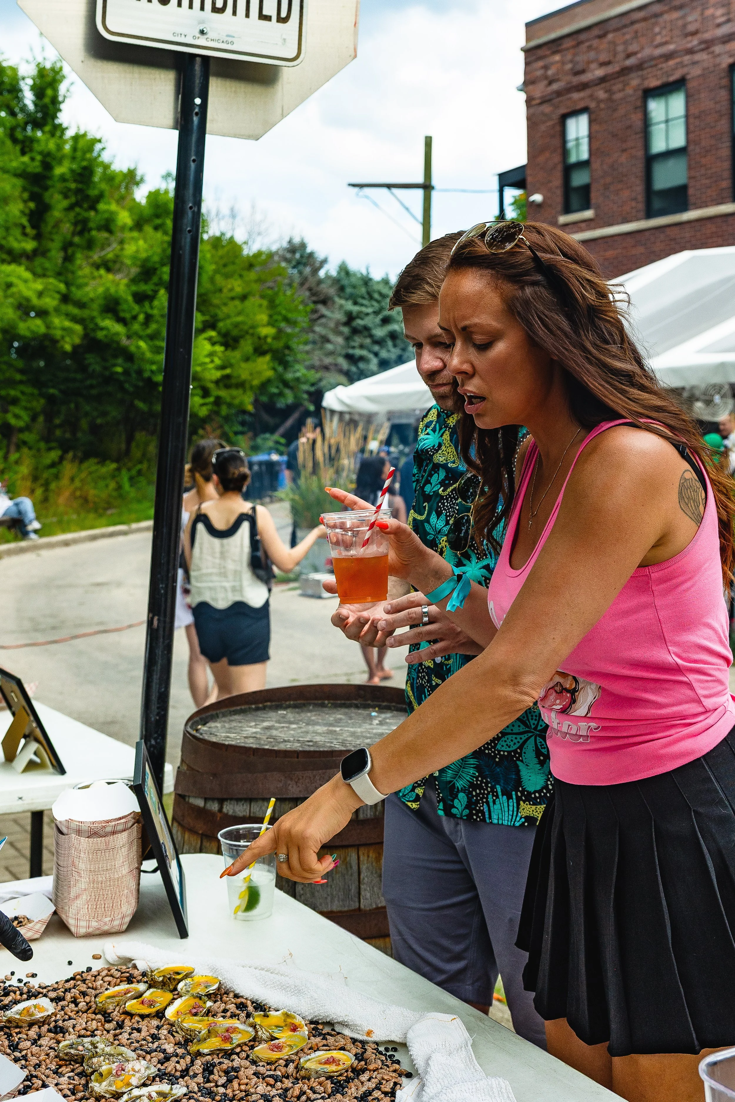 Two women are at an outdoor event looking at a display of food and drinks. One woman in a pink tank top is pointing at the food, while the woman next to her in a colorful patterned shirt is holding a drink with a straw. In the background, there are p