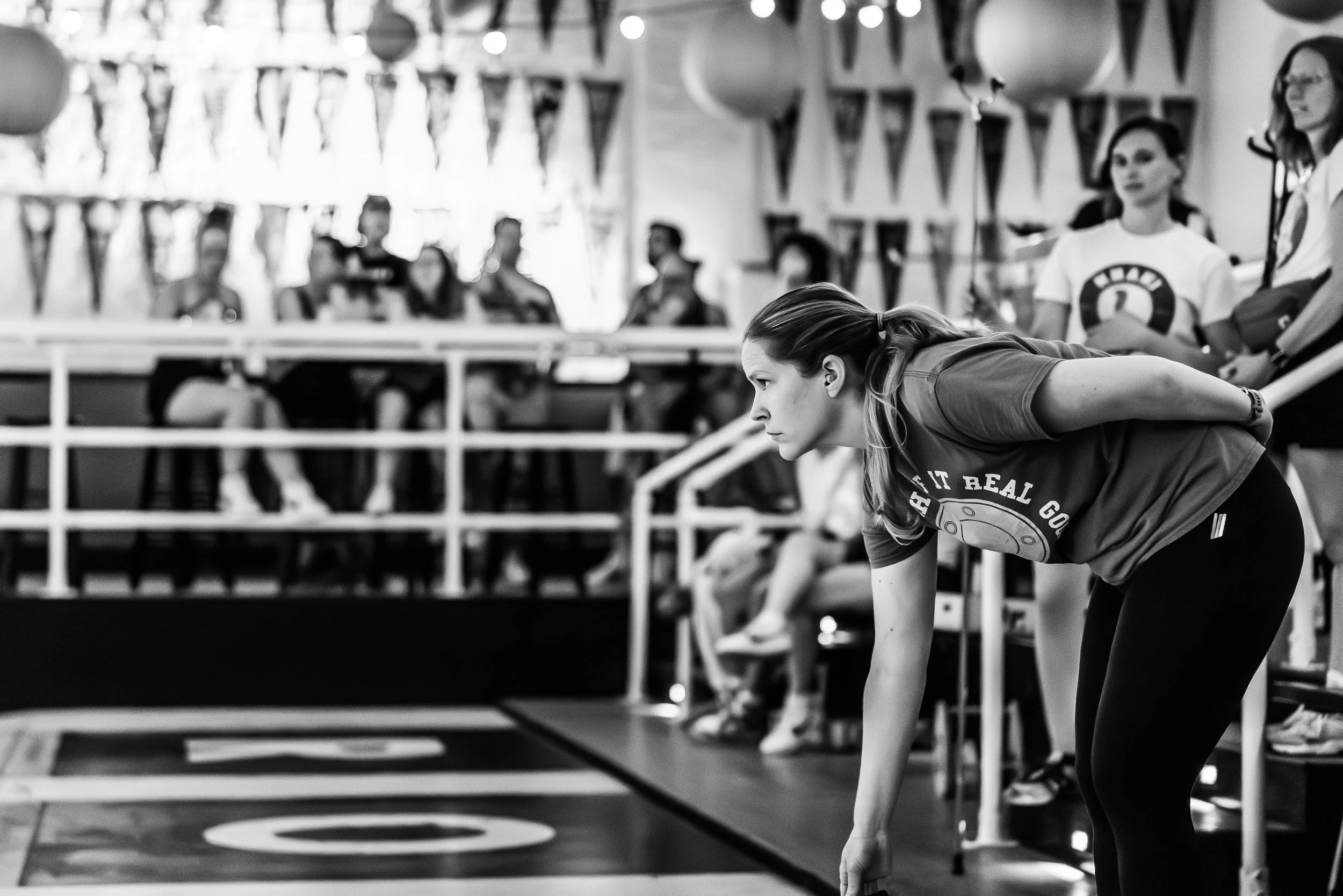 A woman in athletic clothing is in a ready position, leaning forward with her hand on her knee, at a sports event. In the background, several spectators are seated behind a railing, with some wearing jerseys. The setting appears to be an indoor gym o