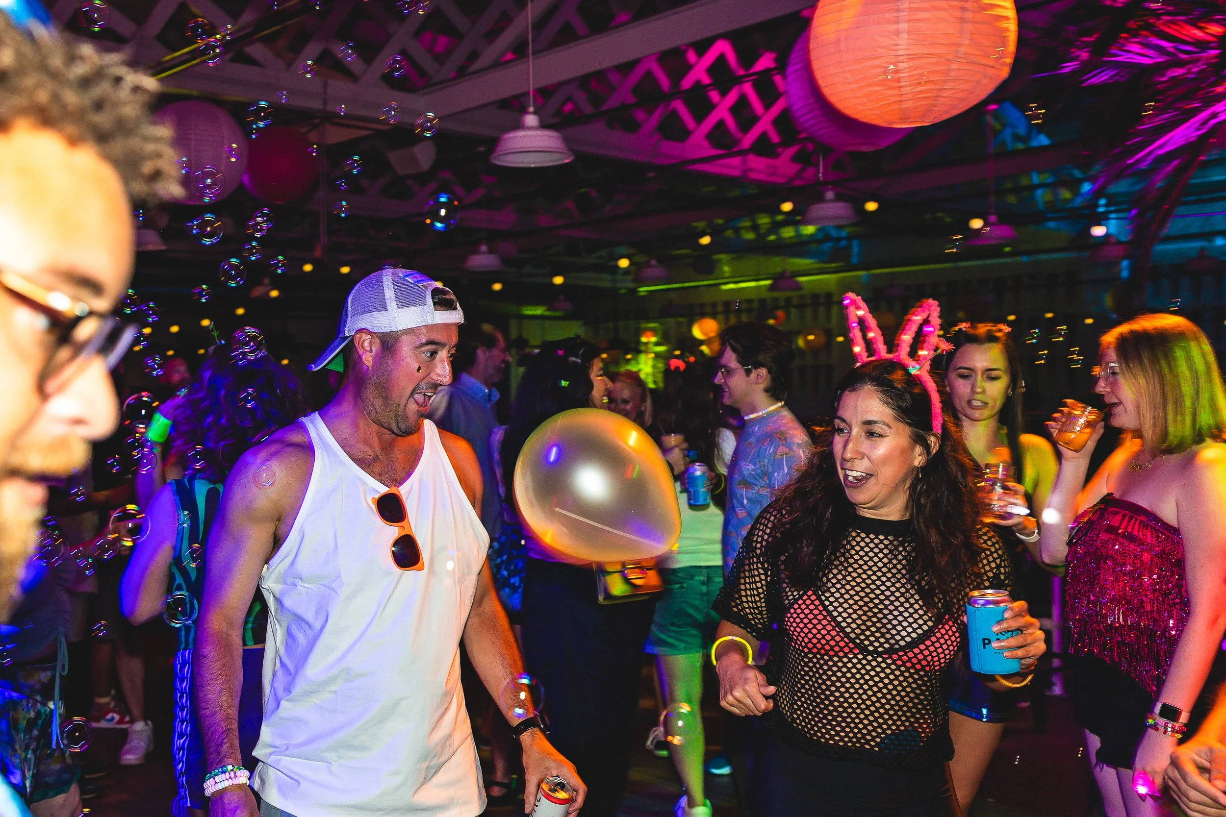 People dancing and celebrating at a lively indoor party with colorful lights, balloons, and bubble effects, some wearing playful accessories like bunny ears.