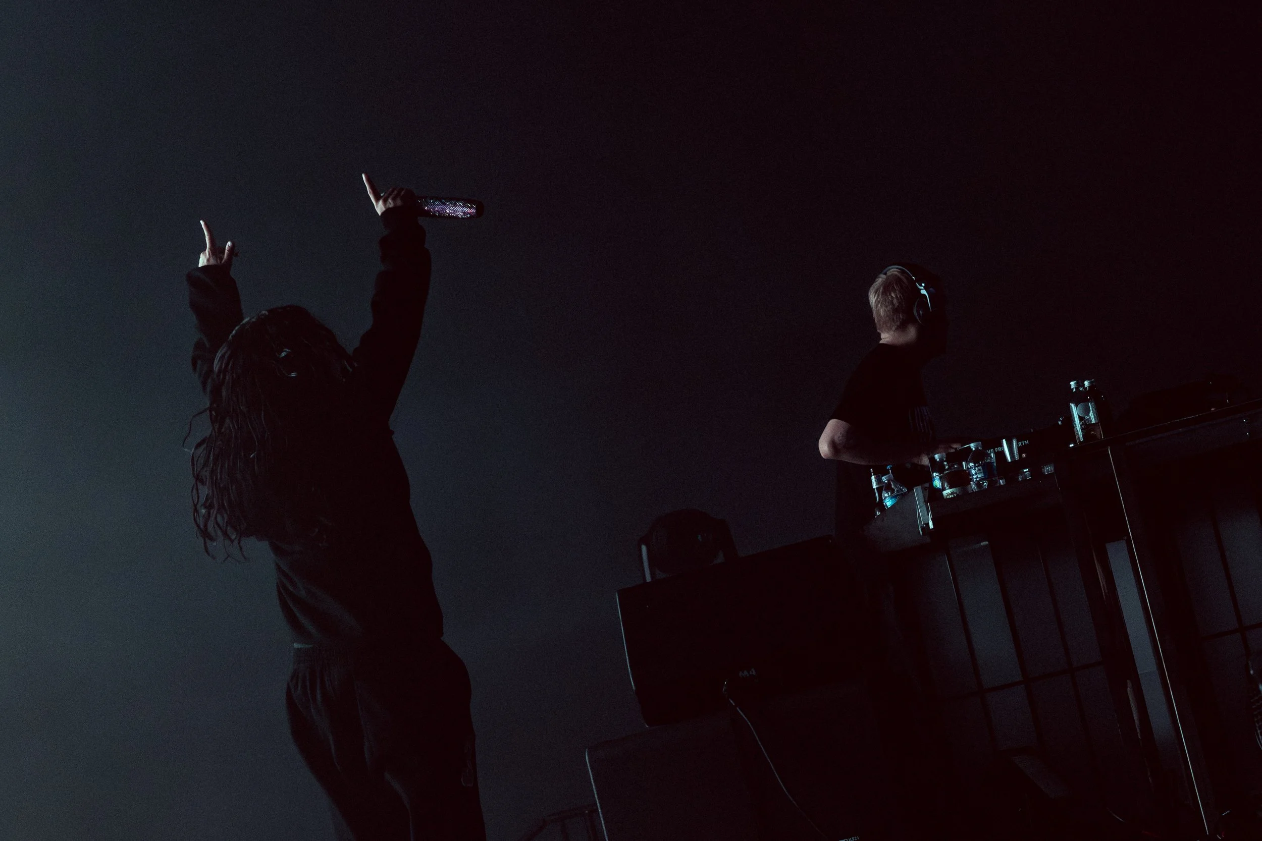 A DJ wearing headphones works at a mixing station with bottles of water, while a woman with curly hair sings or dances with her hands raised, holding a water bottle at a dimly lit event.