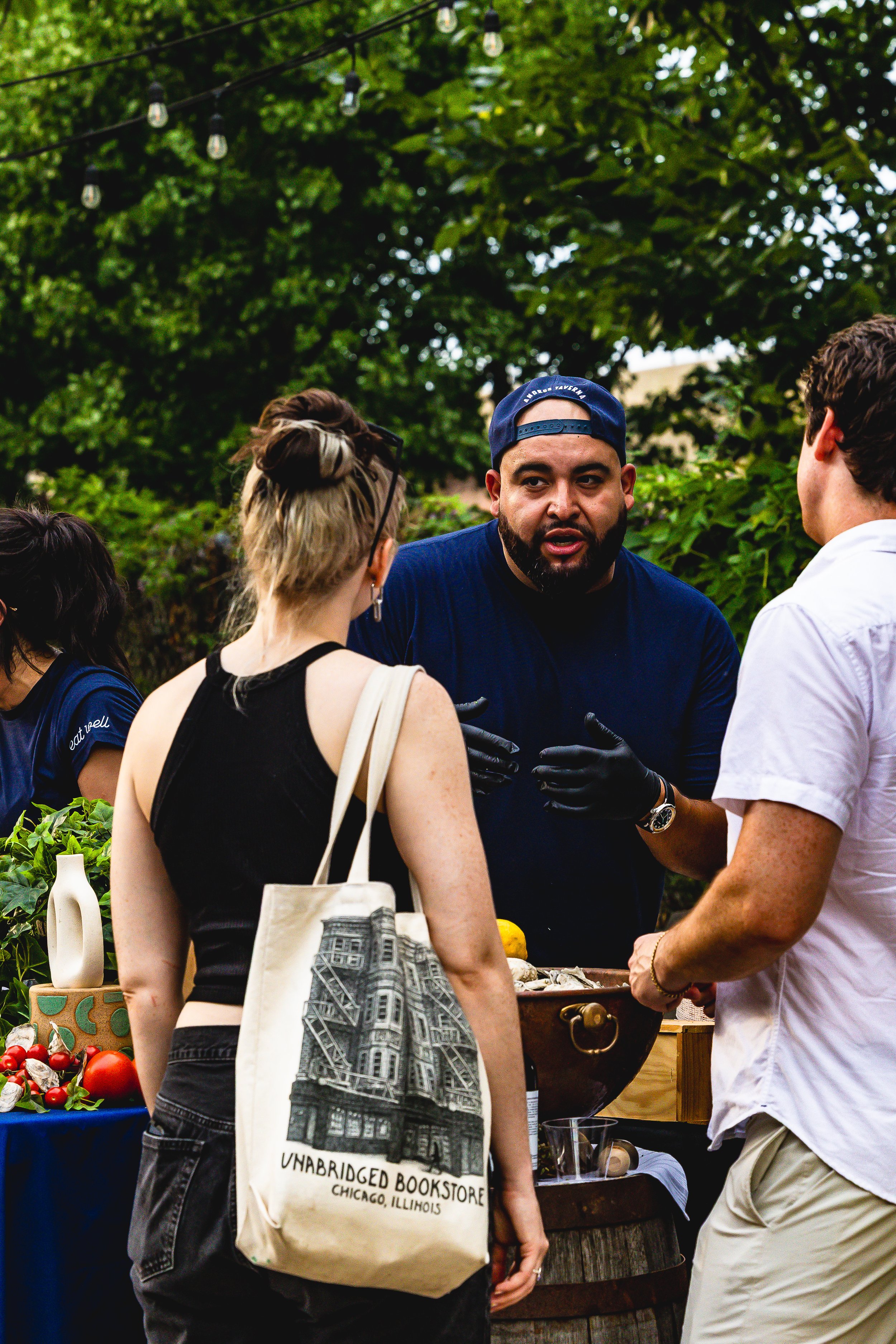 A man with a beard wearing a blue cap and black gloves is talking with two other people at an outdoor event. The woman has a tote bag with a cityscape illustration and the text "Unabridged Bookstore Chicago, Illinois." The background shows green tree