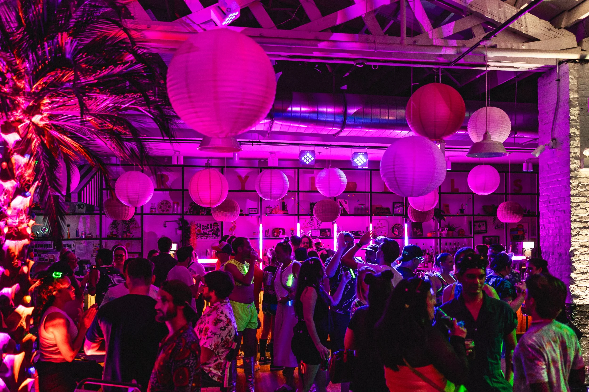 A lively indoor party scene with people dancing and socializing under pink and purple lantern lights. The setting features a neon-lit bar and decorated shelves in the background, with vibrant, colorful lighting creating a festive atmosphere.