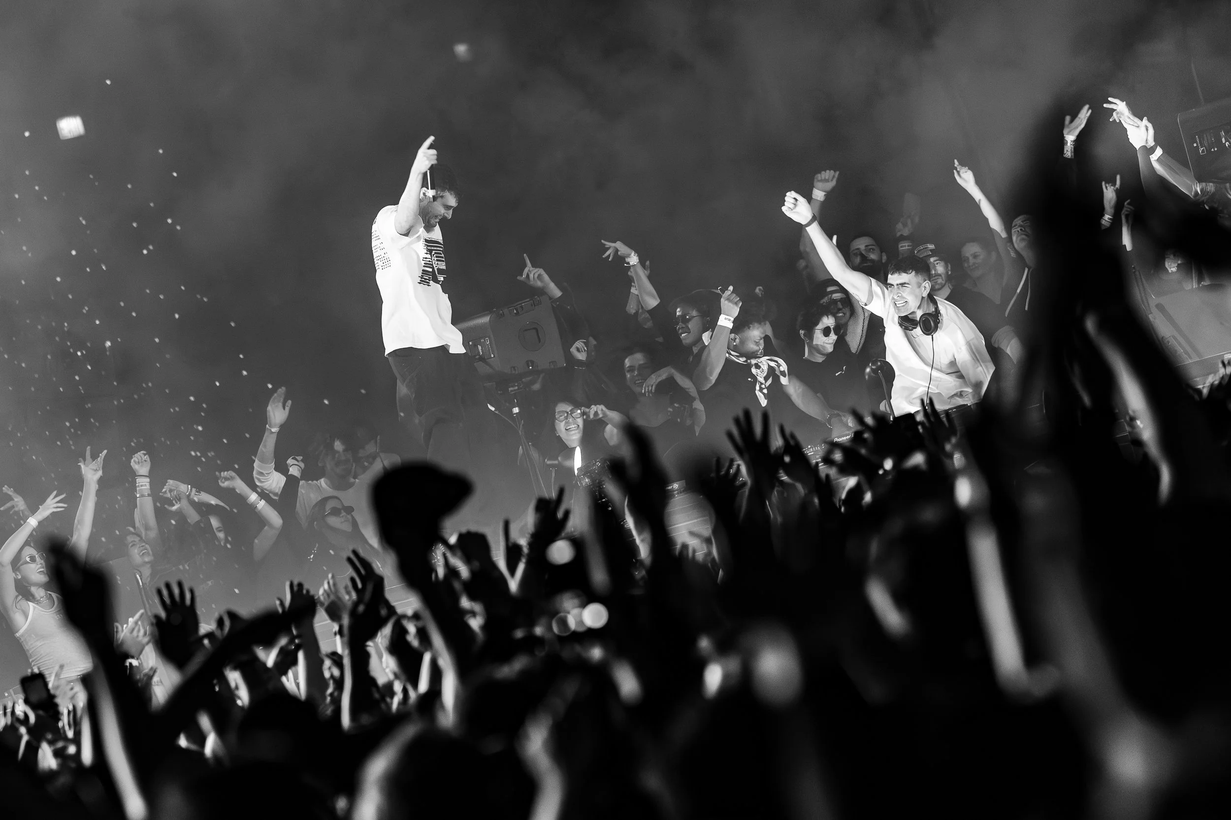 Black and white photo of a lively concert scene with a crowd raising their hands and a Fred Again with headphones on, surrounded by people dancing and enjoying the music.