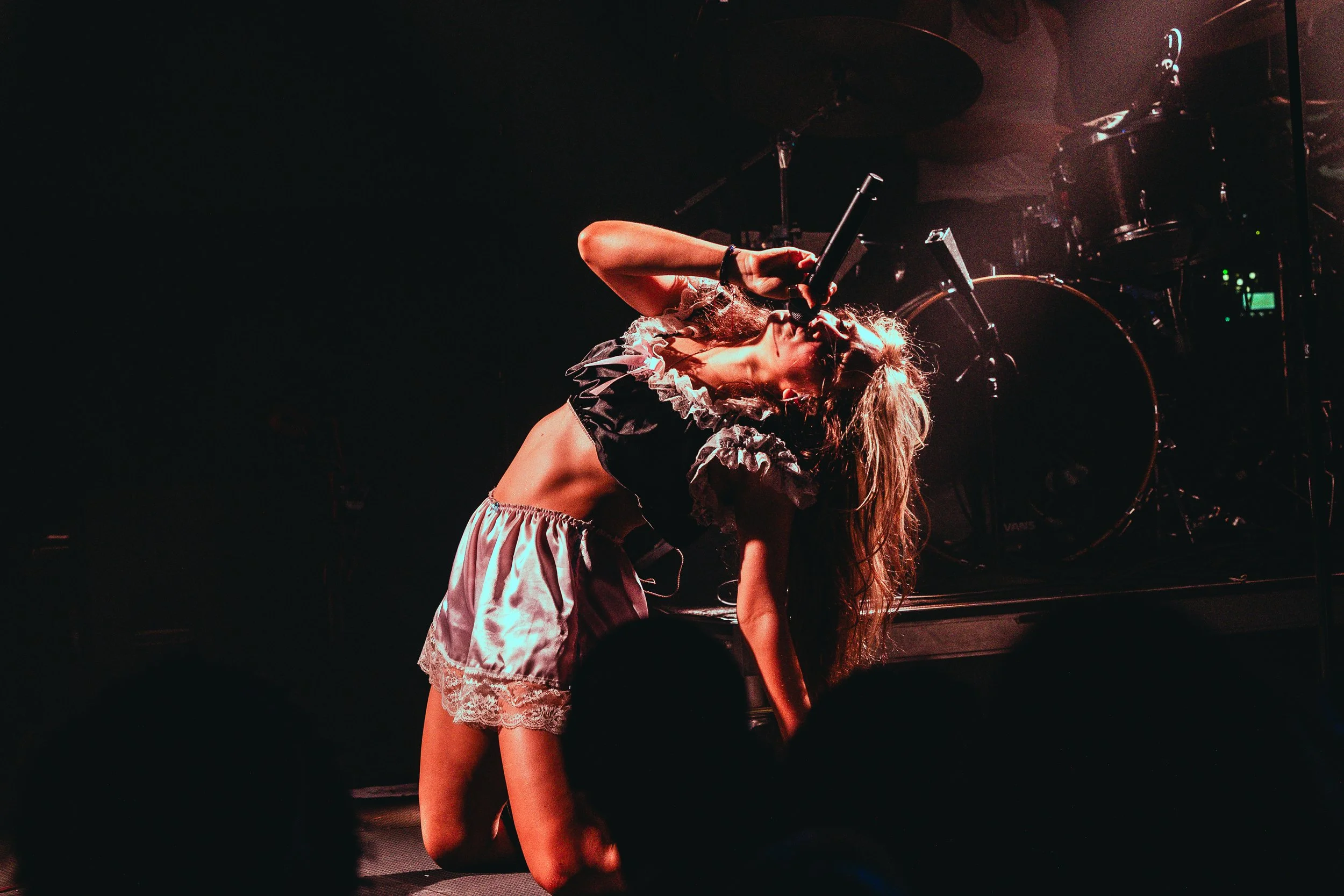 A female singer performing on stage, bent forward, holding a microphone near her mouth, with drums and stage equipment in the background.