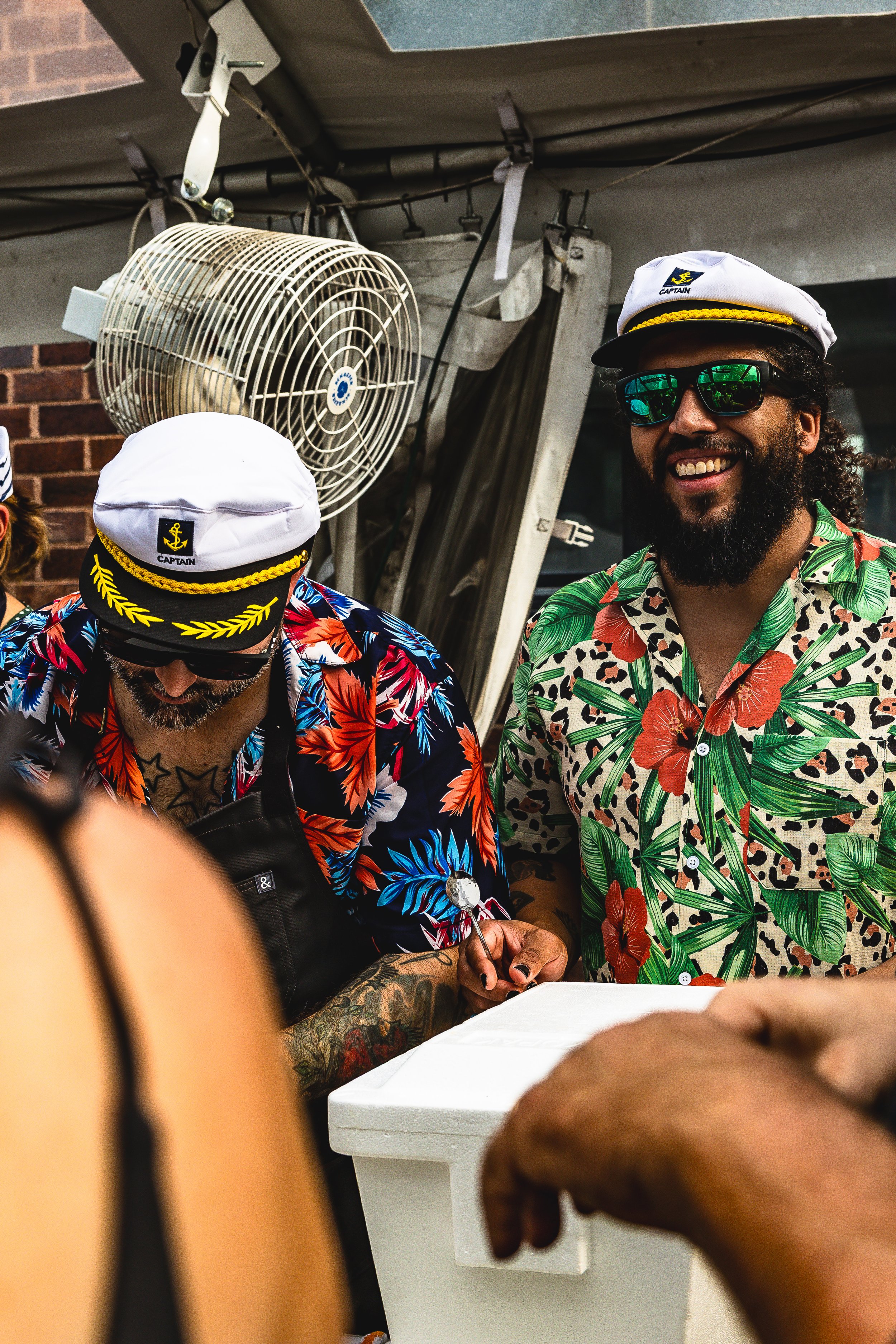 Two men wearing floral Hawaiian shirts, white captain's hats, and sunglasses are smiling and socializing at a beachfront or outdoor setting. There is a fan and some equipment in the background.