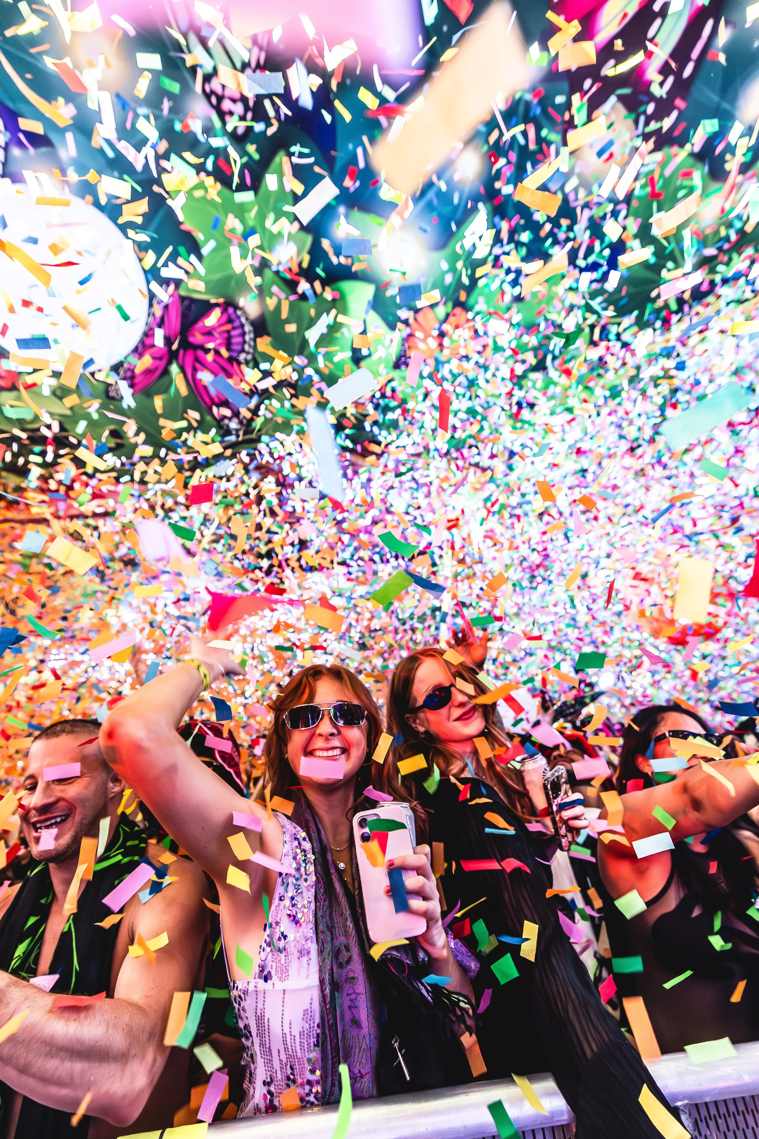 People celebrating at a party with colorful confetti falling from above, wearing sunglasses, smiling, and holding phones.