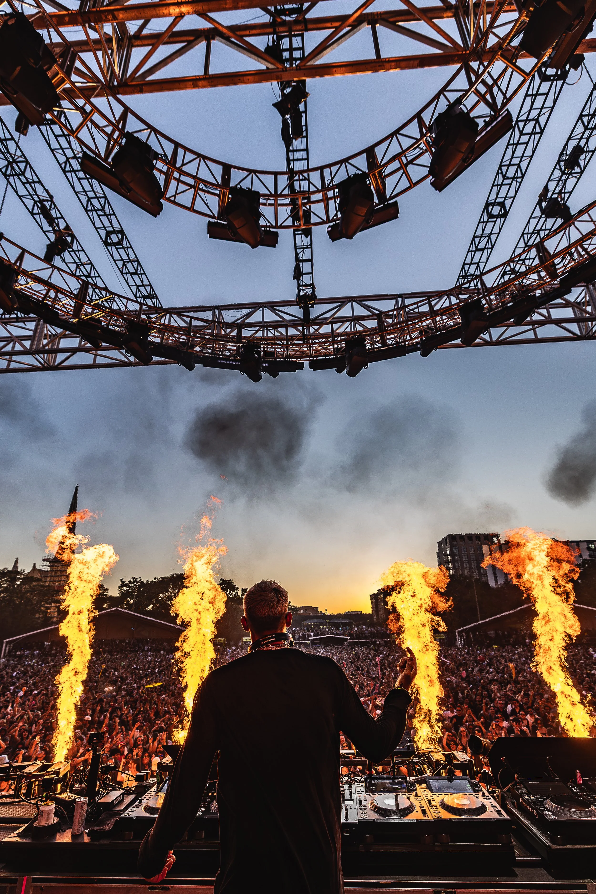 A DJ performing at an outdoor music festival during sunset with pyrotechnics, a large crowd, and stage lighting setup with smoke and fire effects.