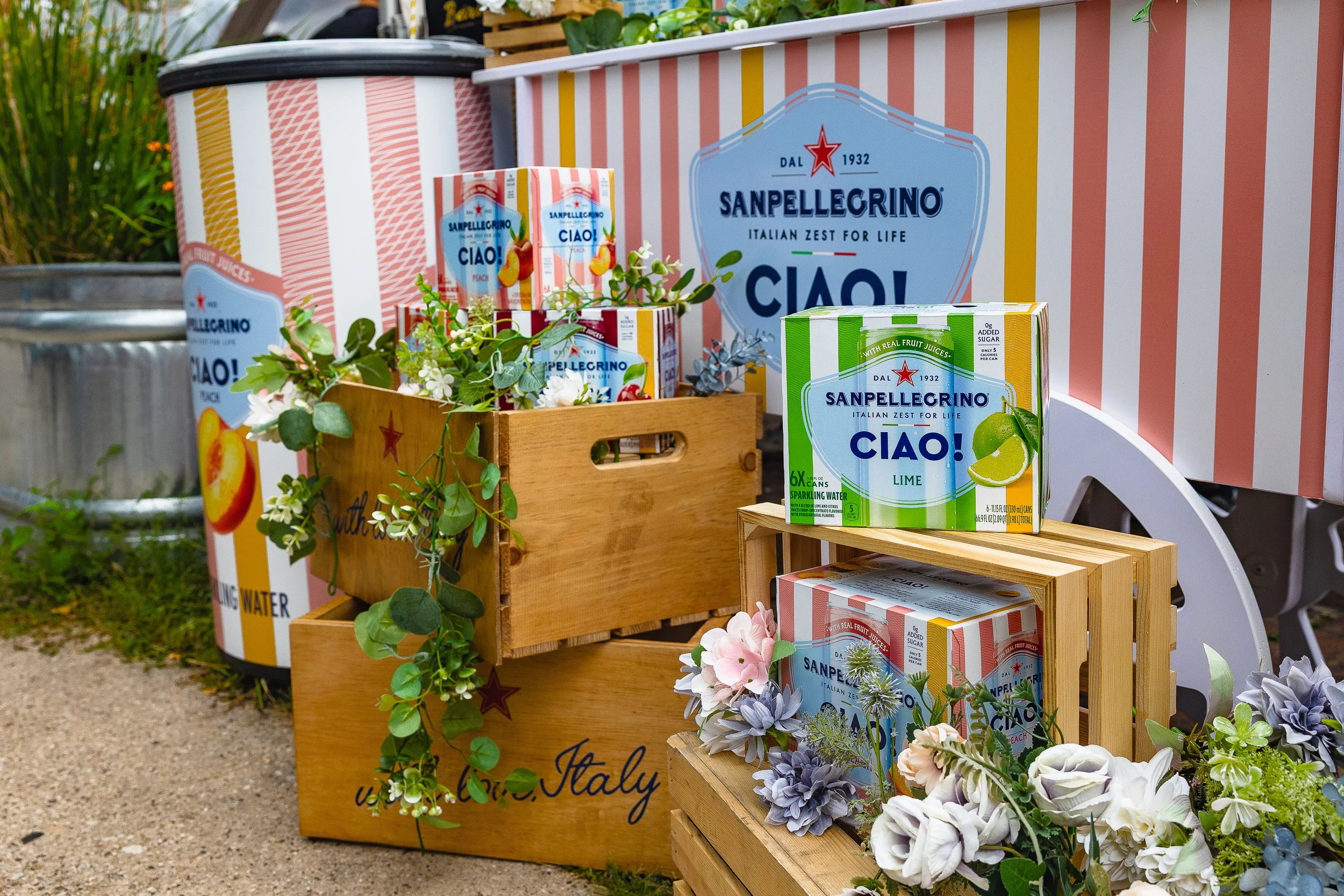 Display of Sanpellegrino beverages, including cans and cartons of lime and peach flavors, arranged with decorative flowers on wooden crates and a large striped box in the background with brand branding.