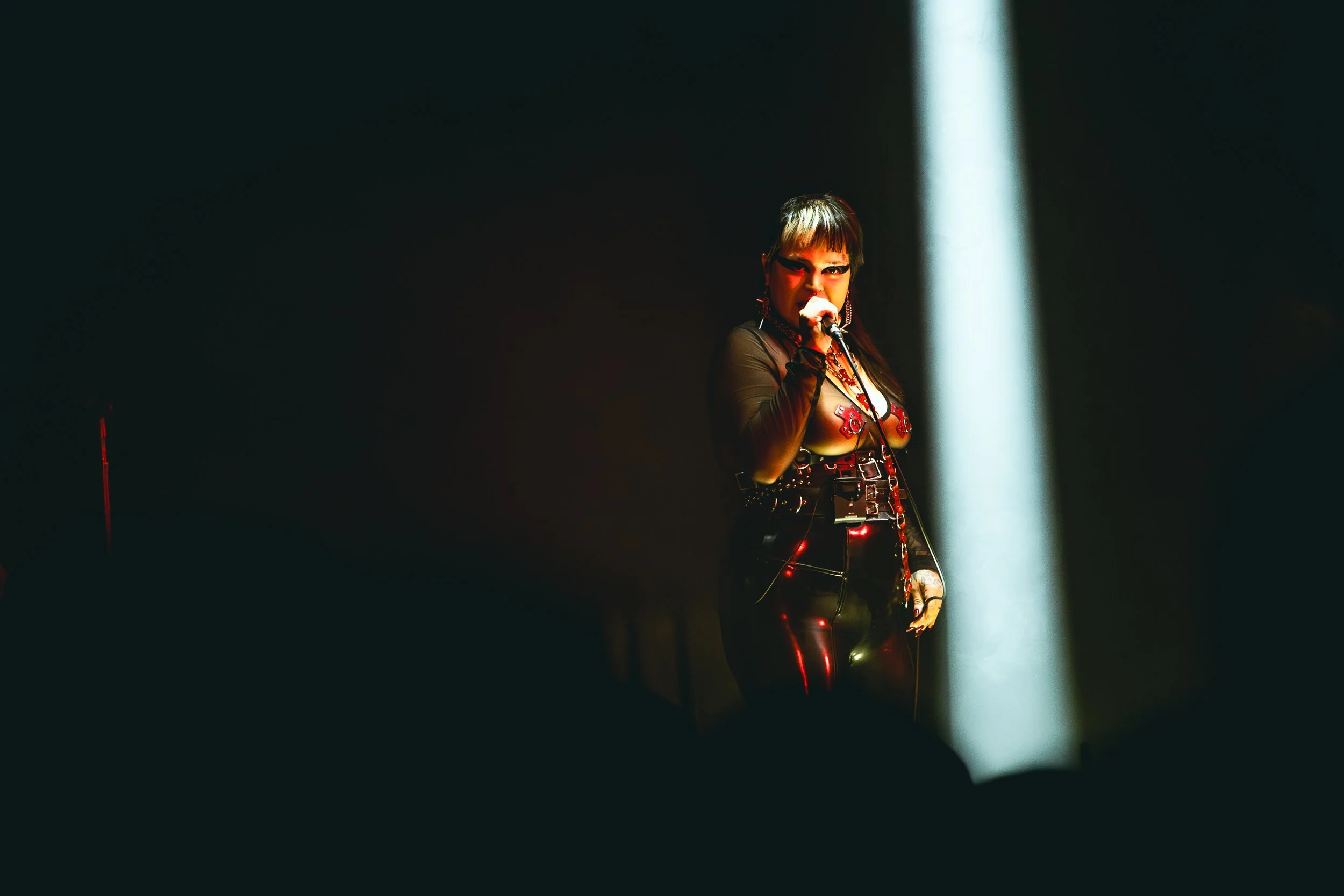 A woman singing on stage with a spotlight on her, wearing a black mesh top with red and black accents, and holding a microphone.