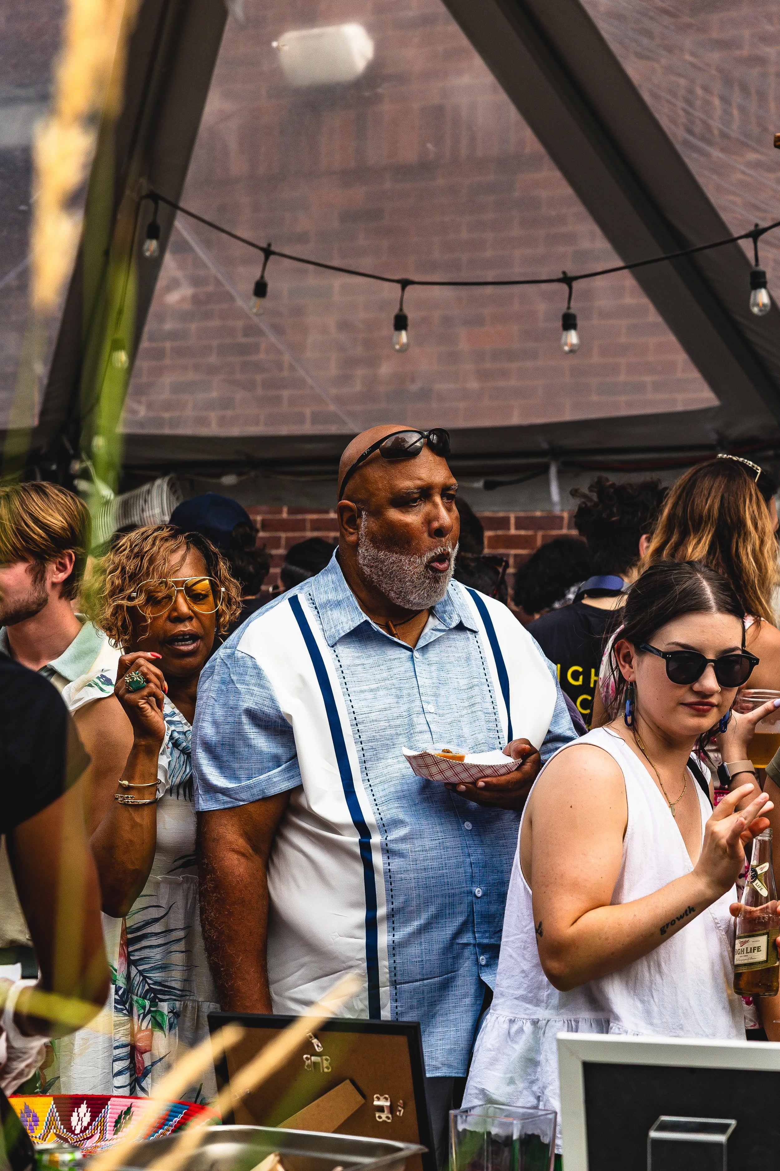A group of people at a social gathering or party in a tent, with a brick wall in the background, hanging string lights, and various displays and drinks.