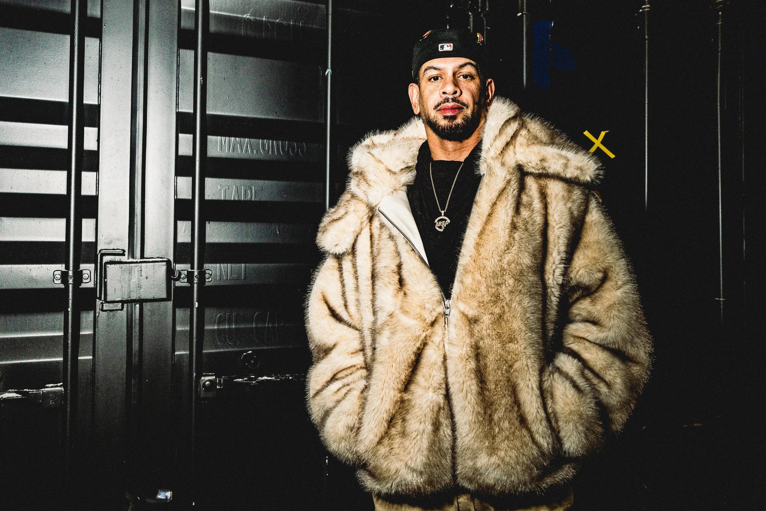 Marc Kinchen, known by his initials MK, wearing a beige fur coat, black baseball cap, and necklaces stands in a dark industrial setting with metallic containers or equipment in the background.