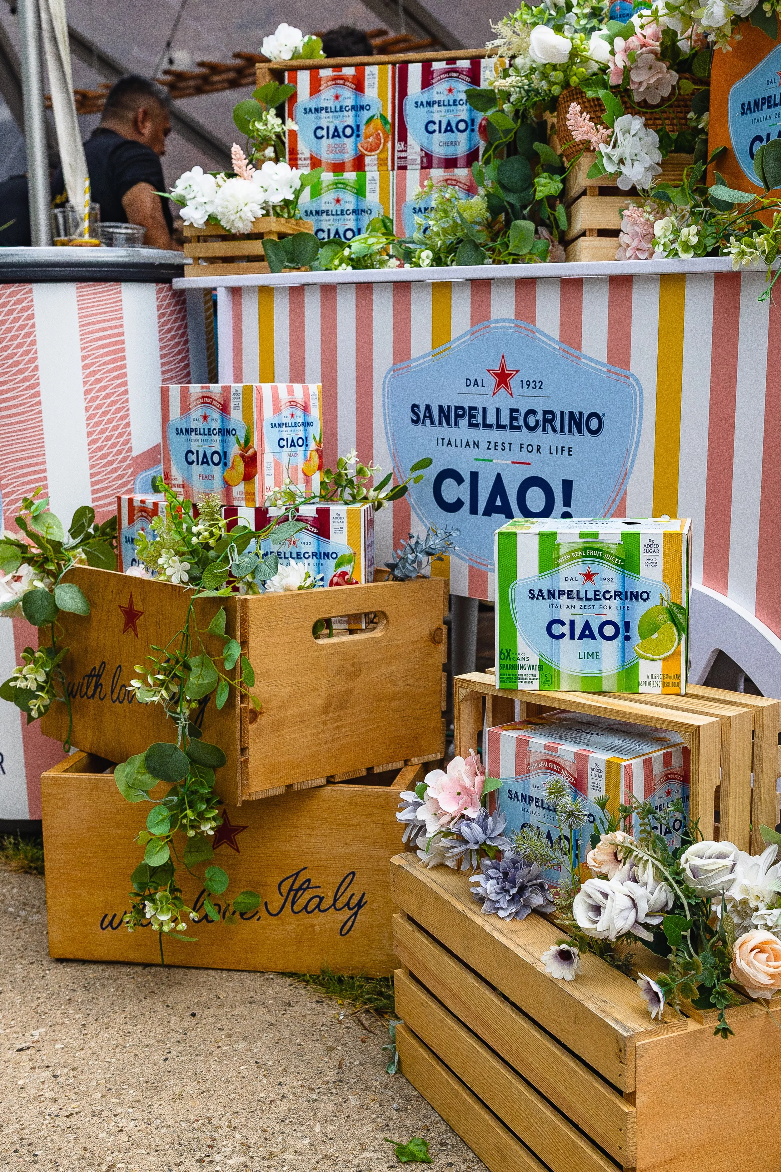 Display of Sanpellegrino canned beverages with floral decorations and wooden crates at a market stall.