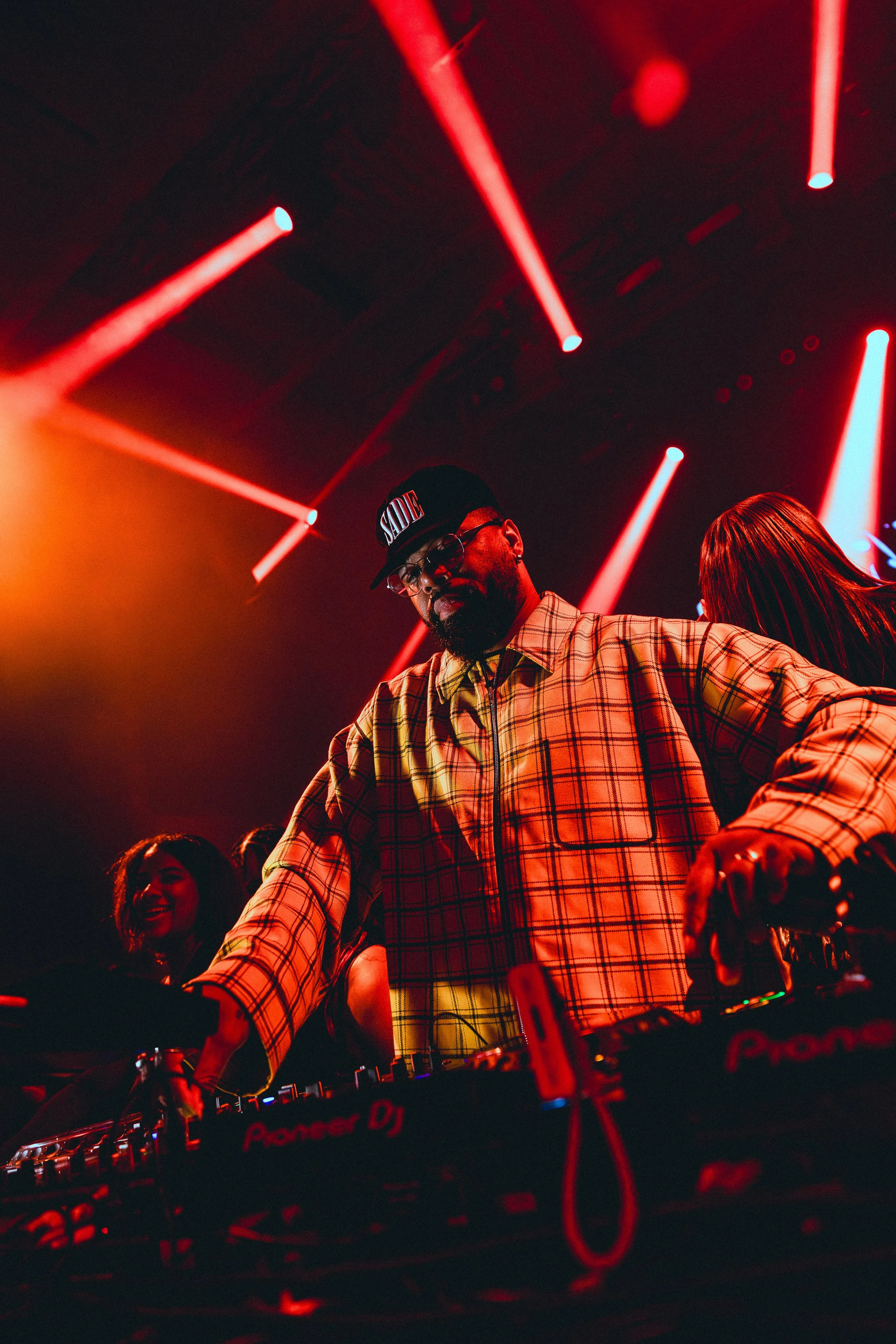 Andre Power wearing a yellow plaid shirt, sunglasses, and a cap labeled 'SADIE' performs at a nightclub with red and blue stage lighting, while smiling women dance behind him.