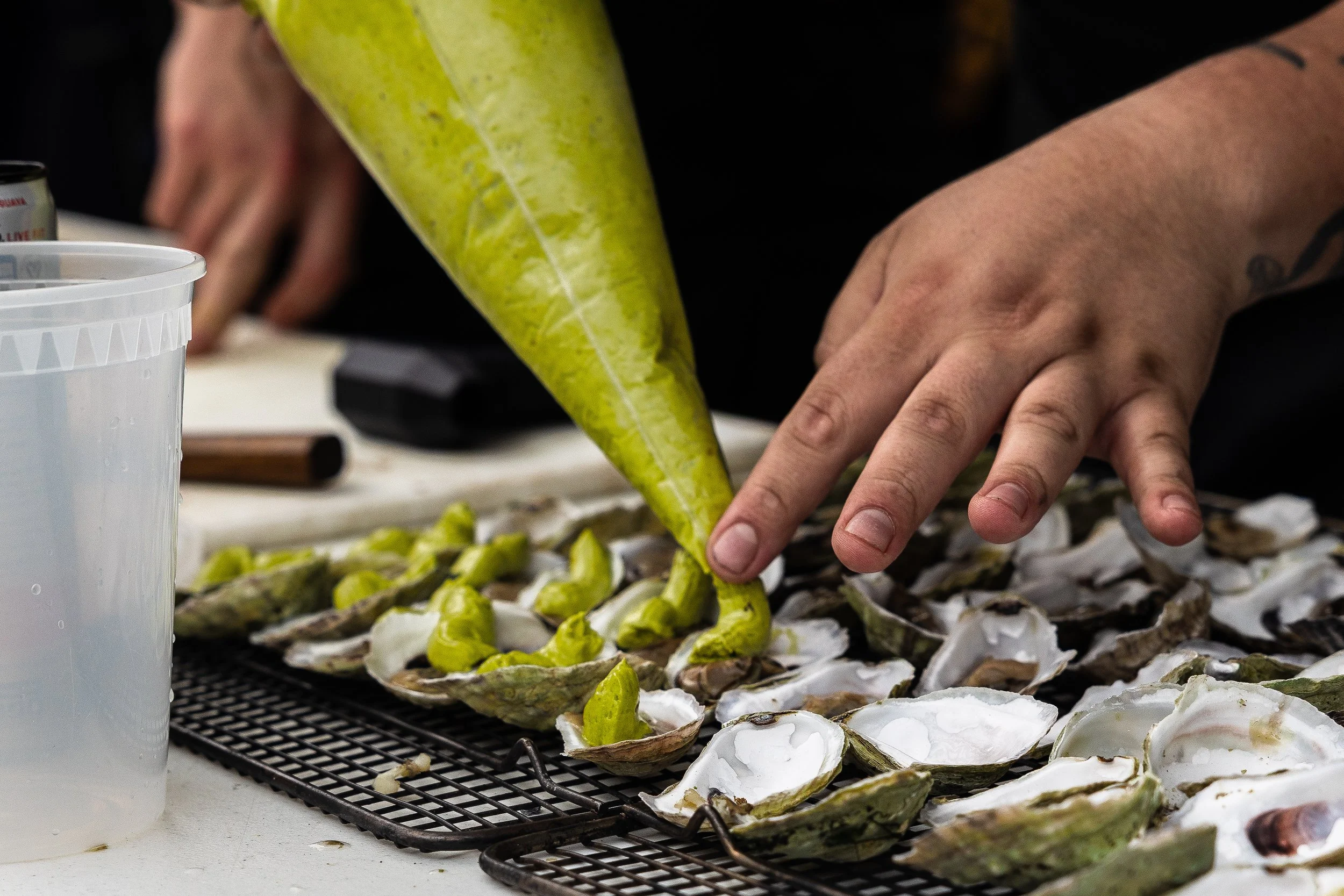 Close-up of a person's hand shucking oysters on a wire rack, with green oysters and shells on a white surface.