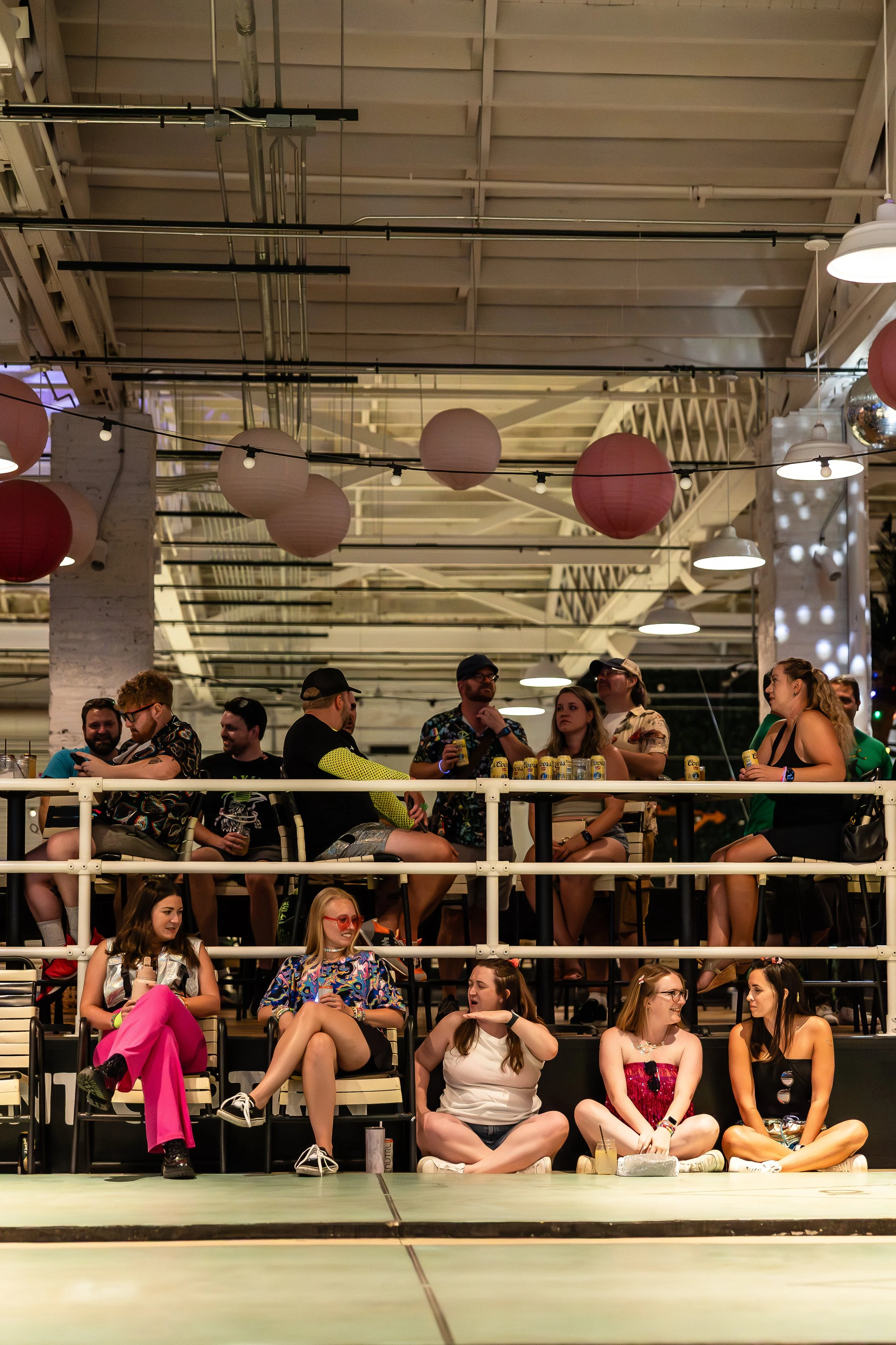 Group of people socializing in a lively indoor space decorated with pink and white paper lanterns, some sitting on chairs and others on the floor, with drinks in hand.