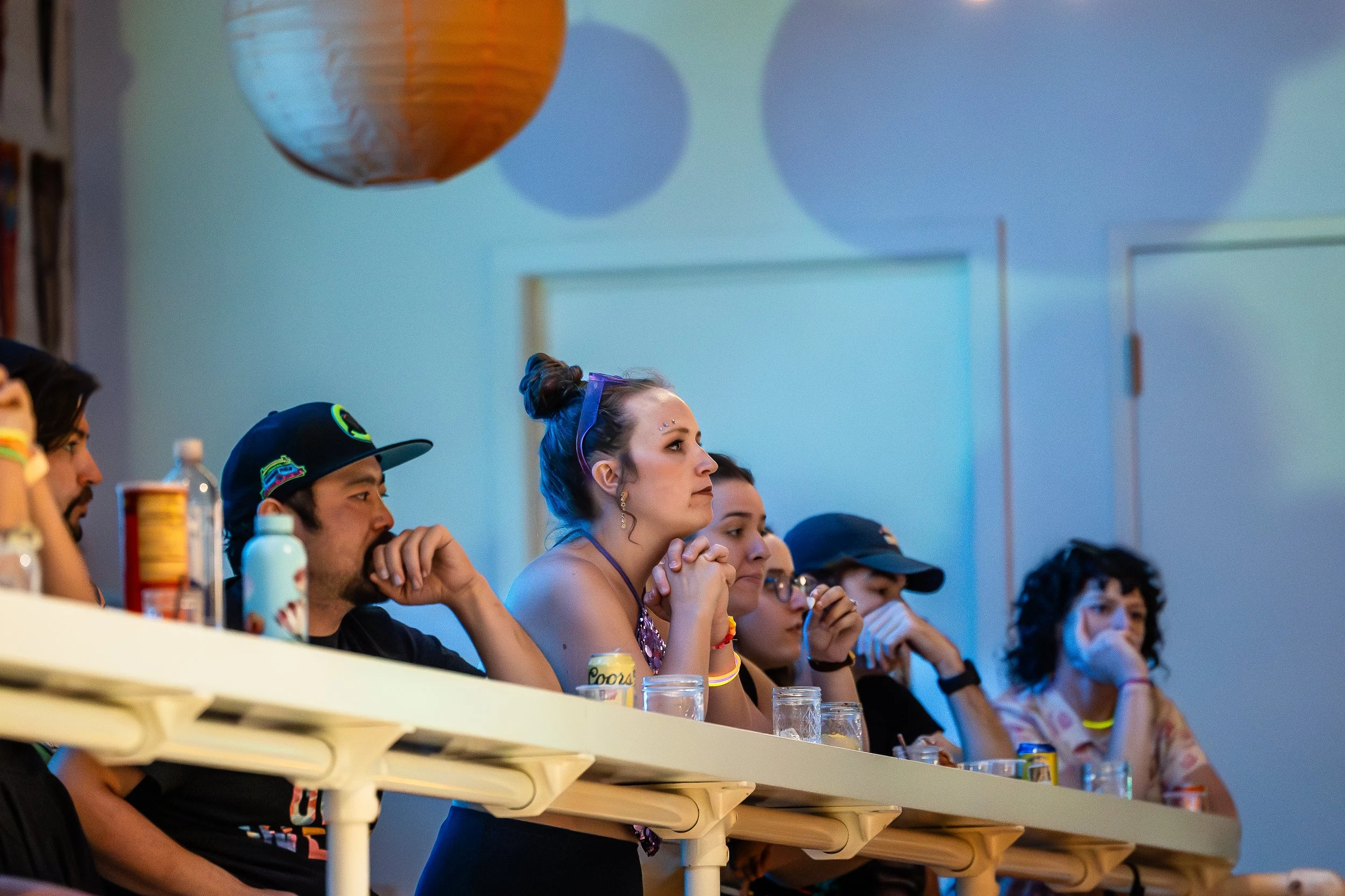 Group of people sitting at a table, watching an event attentively, in a room with blue lighting and paper lanterns on the ceiling.