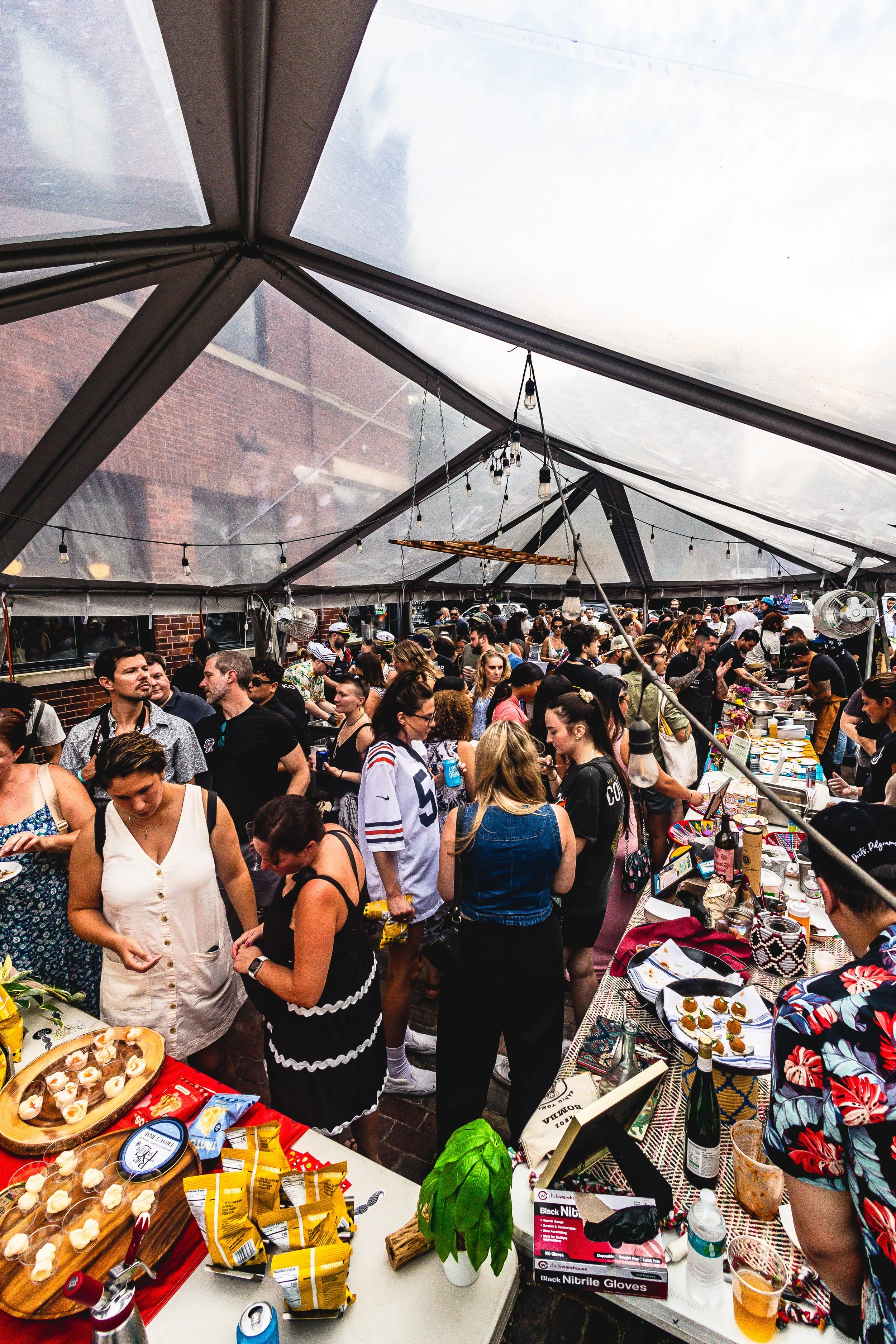 Crowd of people at a lively indoor or outdoor event, with food and drinks on tables, under a large transparent tent.