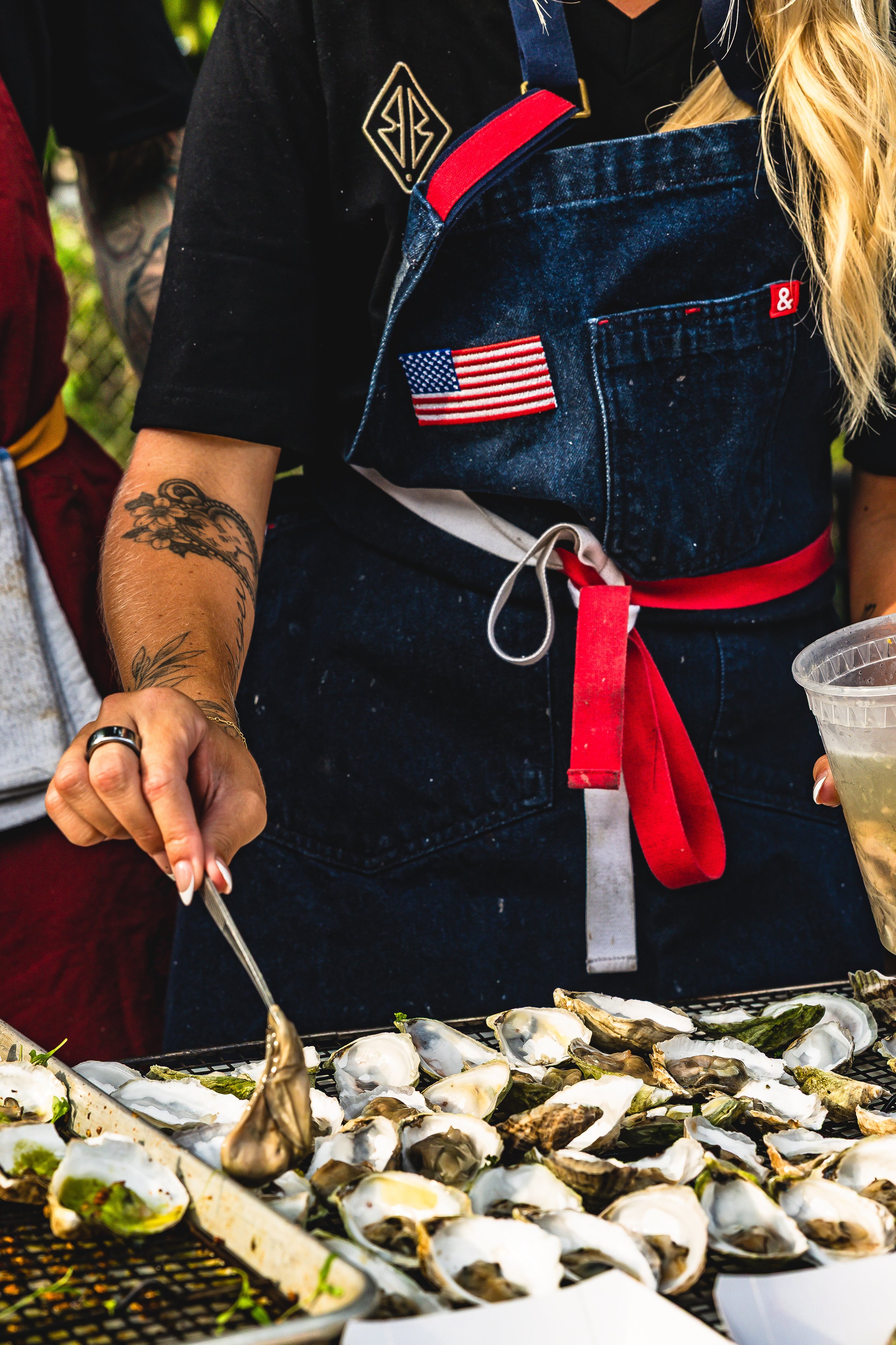 Person shucking oysters with a tray of oysters on a grill, wearing a black T-shirt, denim apron with American flag patch, and red apron ties, outdoors.