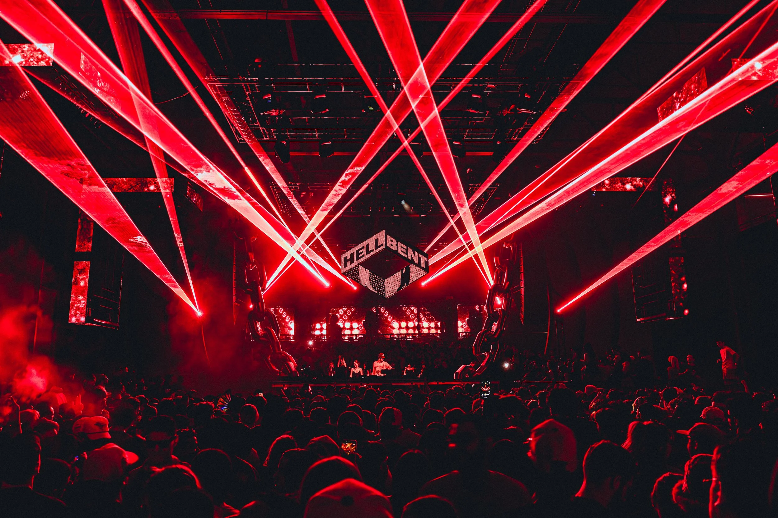 A concert stage with bright red laser lights and a large cube-shaped screen displaying the word 'HELLOBENT' amid an audience.
