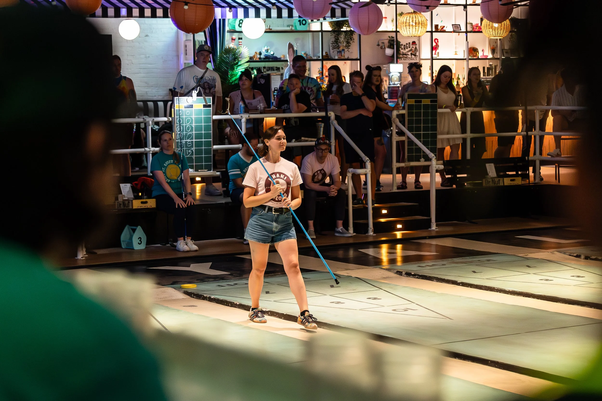 A young woman in casual attire, including a white t-shirt and denim shorts, is playing shuffleboard indoors. She is holding a shuffleboard cue, preparing to slide a disc on the court. Behind her, an audience of people, some seated and some standing, 