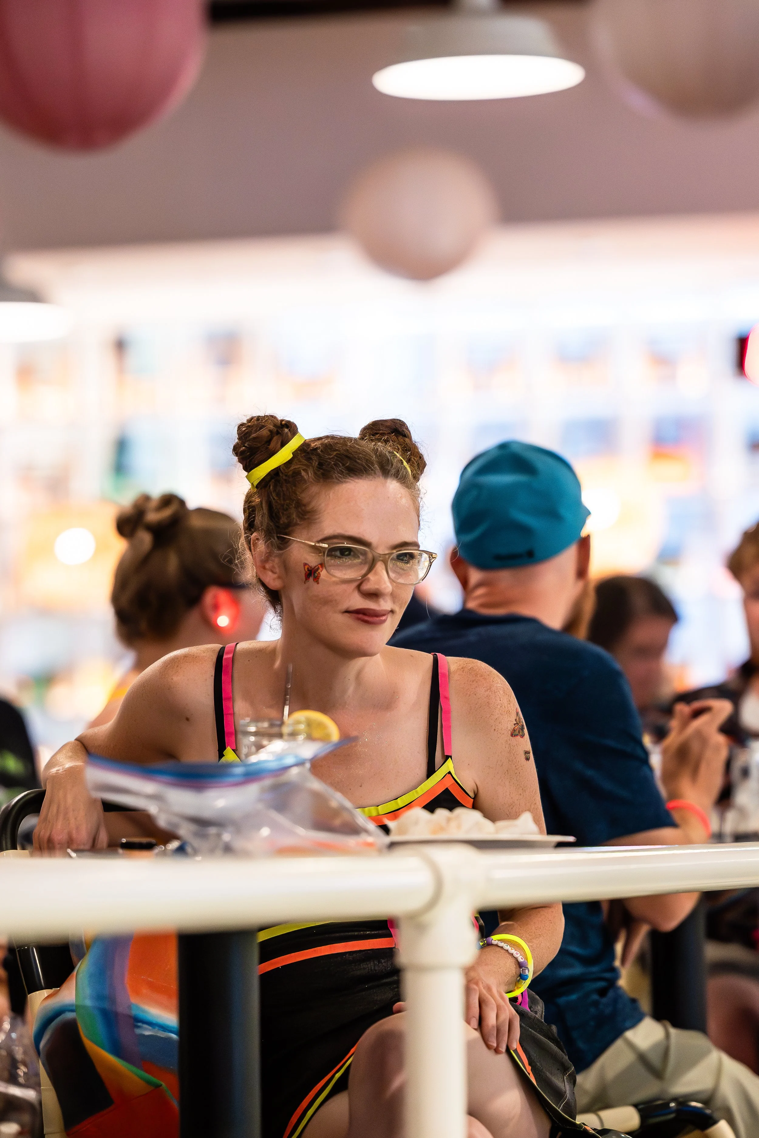 A woman with glasses and colorful hair in buns, wearing a black dress with rainbow accents, sitting at a table with a drink and snacks, in a lively indoor setting with balloons overhead and a brightly lit background.