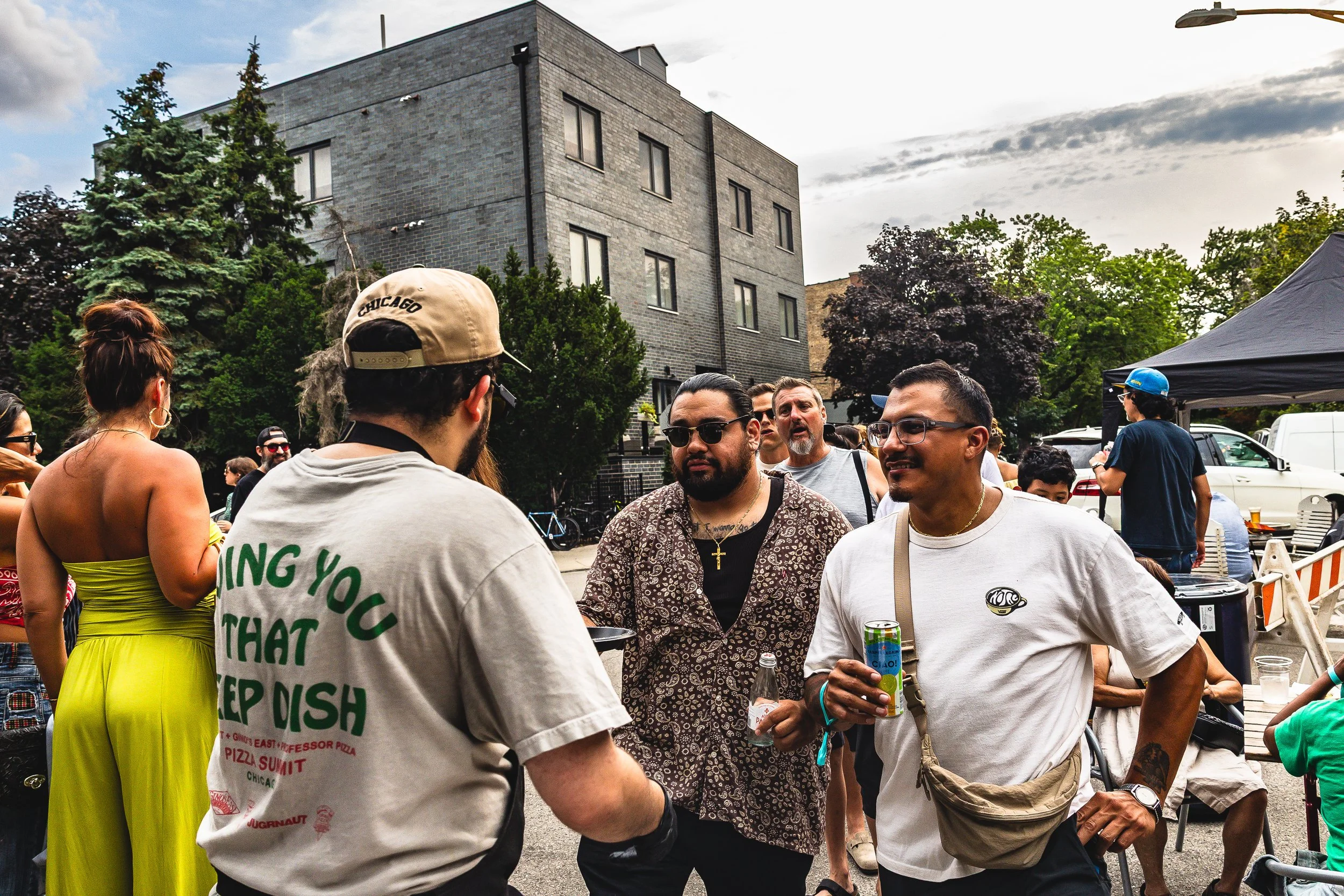 A group of people talking at an outdoor event on a city street, with trees, buildings, and a tent in the background.
