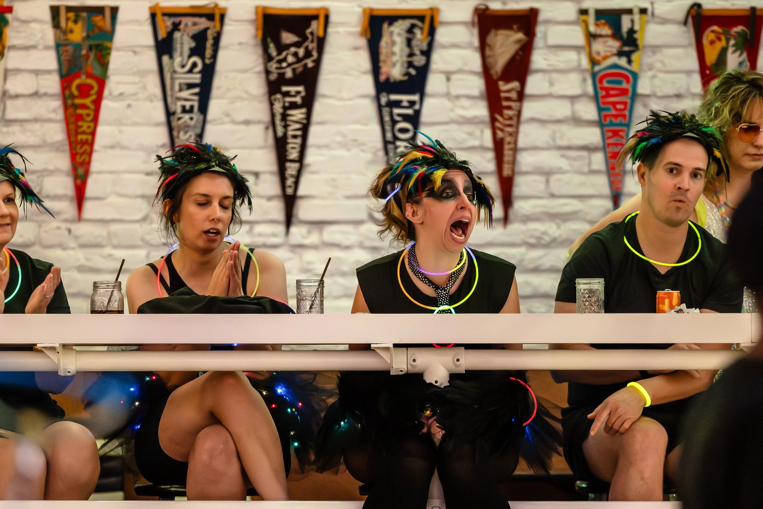 People sitting at a party table wearing colorful feathered headbands and glow necklaces, with banners in the background reading 'Cypress,' 'West Avenue,' 'Floyd,' 'Jersey,' and 'CAPE FEAR.' The woman in the middle is yelling or shouting, while others
