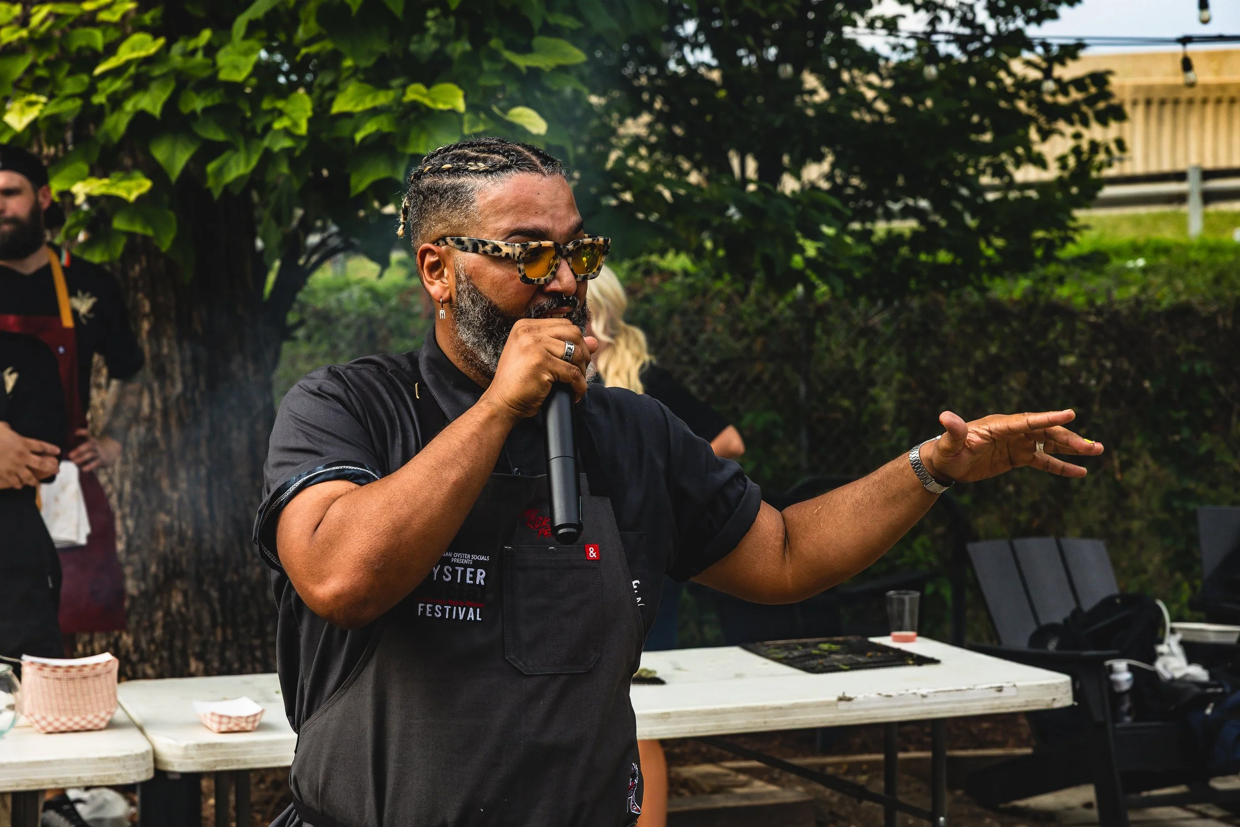 A man wearing sunglasses and a black apron speaking into a microphone at an outdoor event.