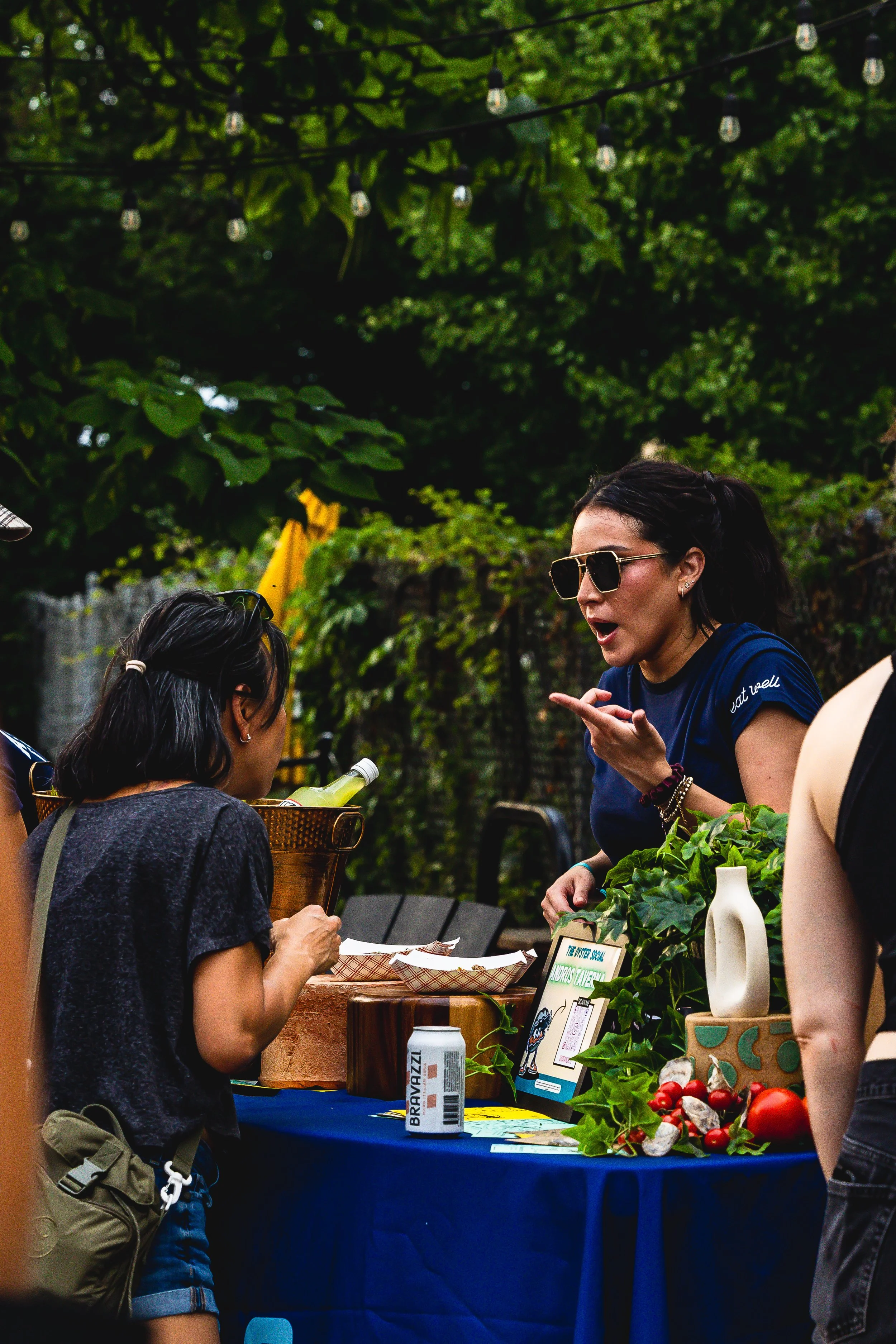 A woman with dark hair and sunglasses is talking to a girl at a table decorated with plants and tomatoes, outdoors with green trees and string lights overhead.