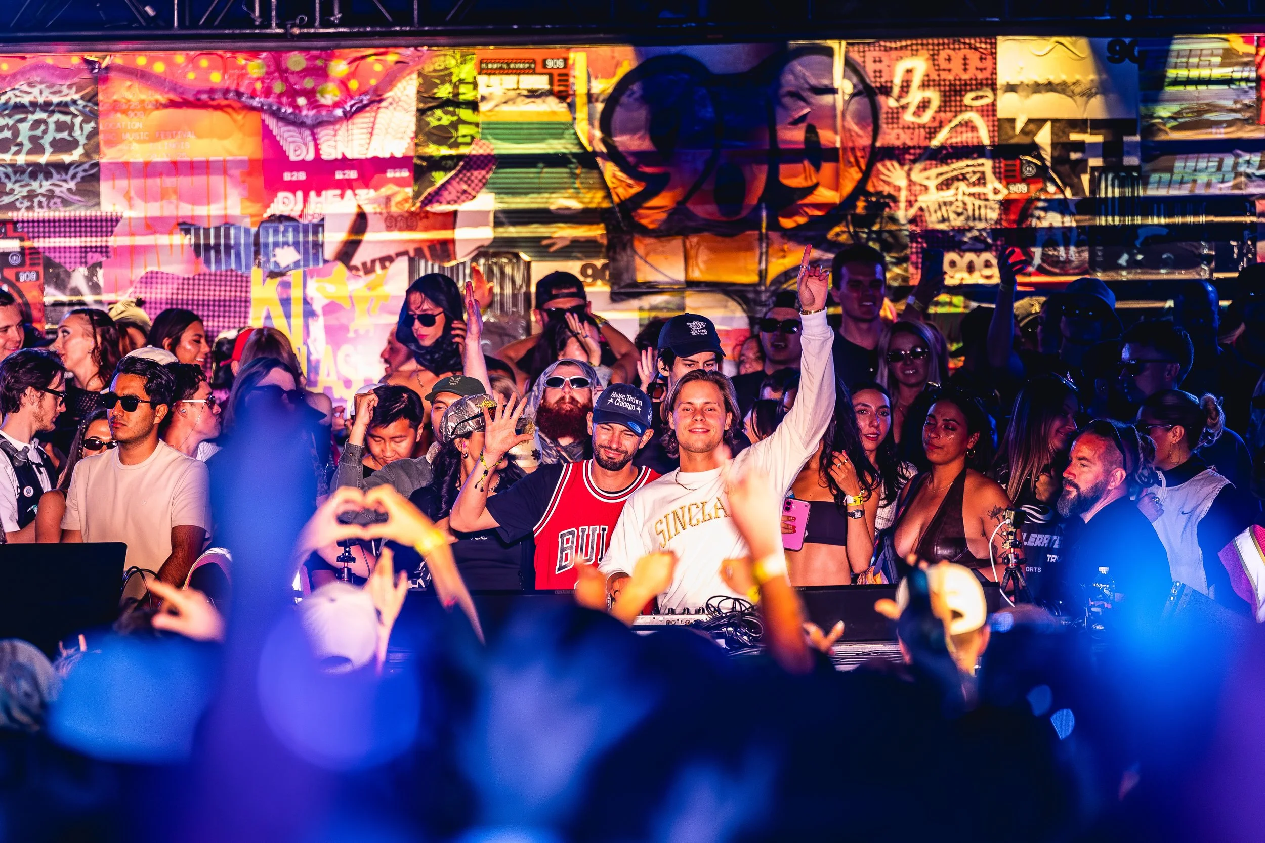 Crowd of diverse people at a music festival or concert, with two men in the front raising their hands, surrounded by others enjoying the event, colorful graffiti art in the background.